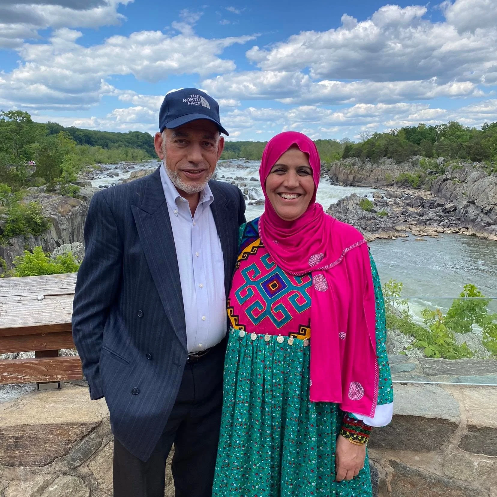 A man and a woman stand together outdoors by a river, smiling at the camera on a partly cloudy day with green trees in the background.