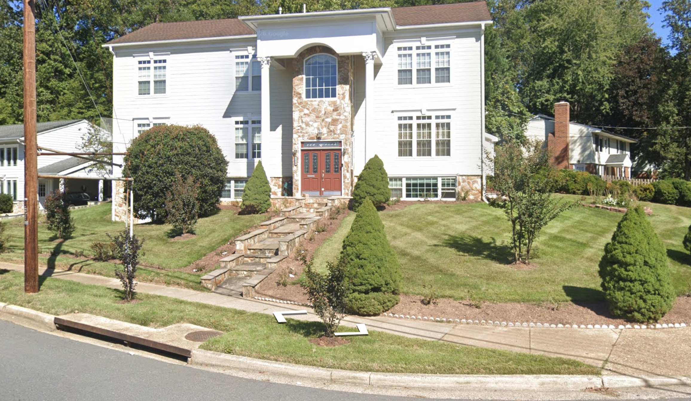 A two-story white house with a stone accent wall and a red front door, surrounded by a landscaped lawn with small trees and bushes, and a sidewalk in the foreground.