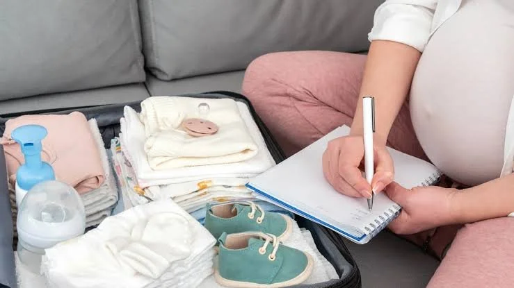 A person sitting on a pink couch with an open suitcase filled with baby items, including diapers, clothes, a pacifier, diapers cream, baby shoes, and bottles. The person is writing in a notebook with a pen.