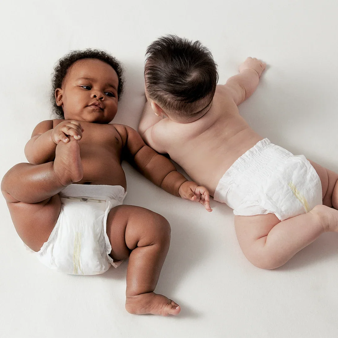 Two babies lying on a white surface, one with darker skin and curly hair, the other with lighter skin and straight hair, both wearing diapers.