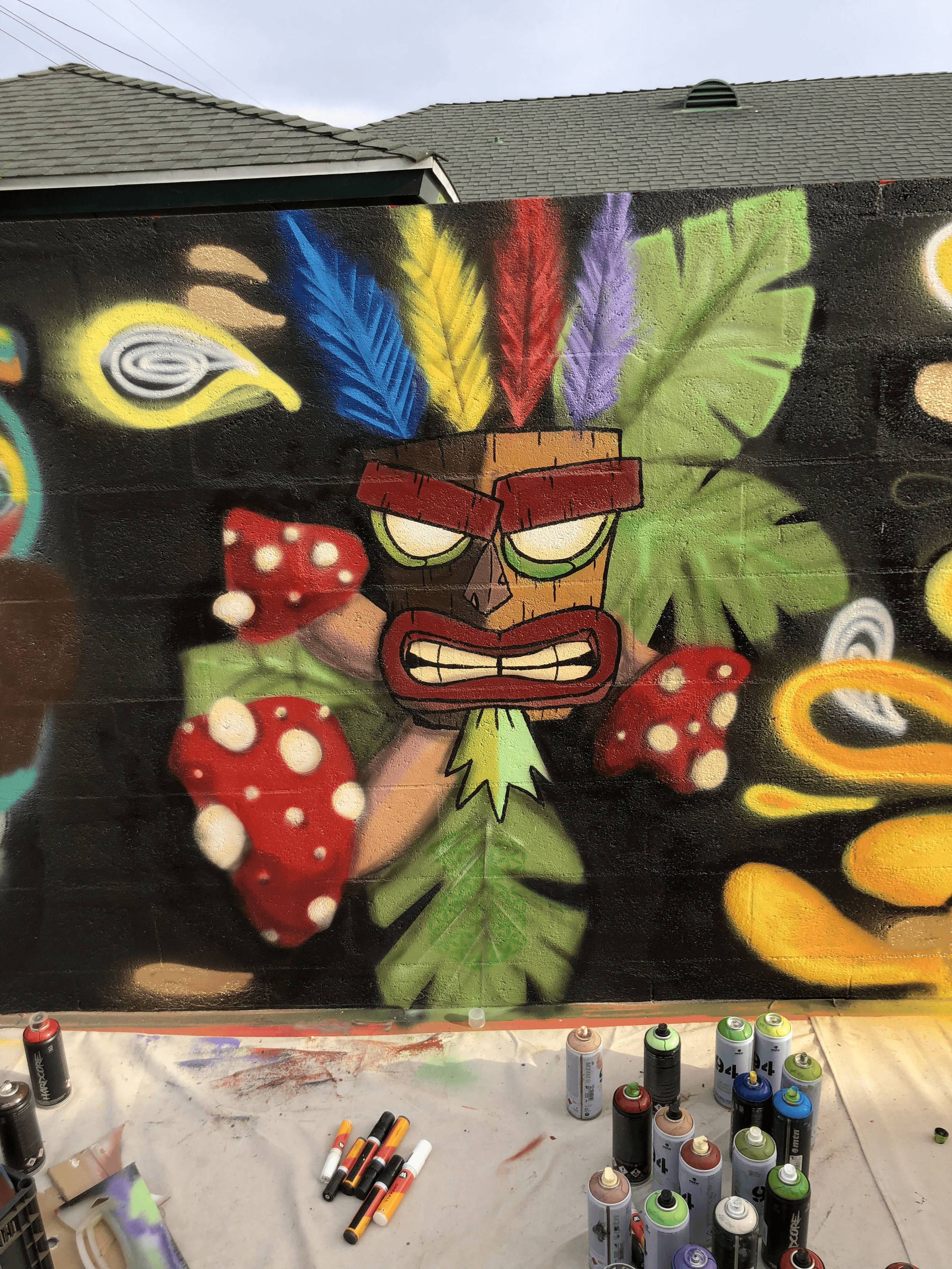 Graffiti art featuring ApuApu mask from 90's videogame Crash Bandicoot, surrounded by colorful leaves and abstract shapes, with spray paint cans on the table below.