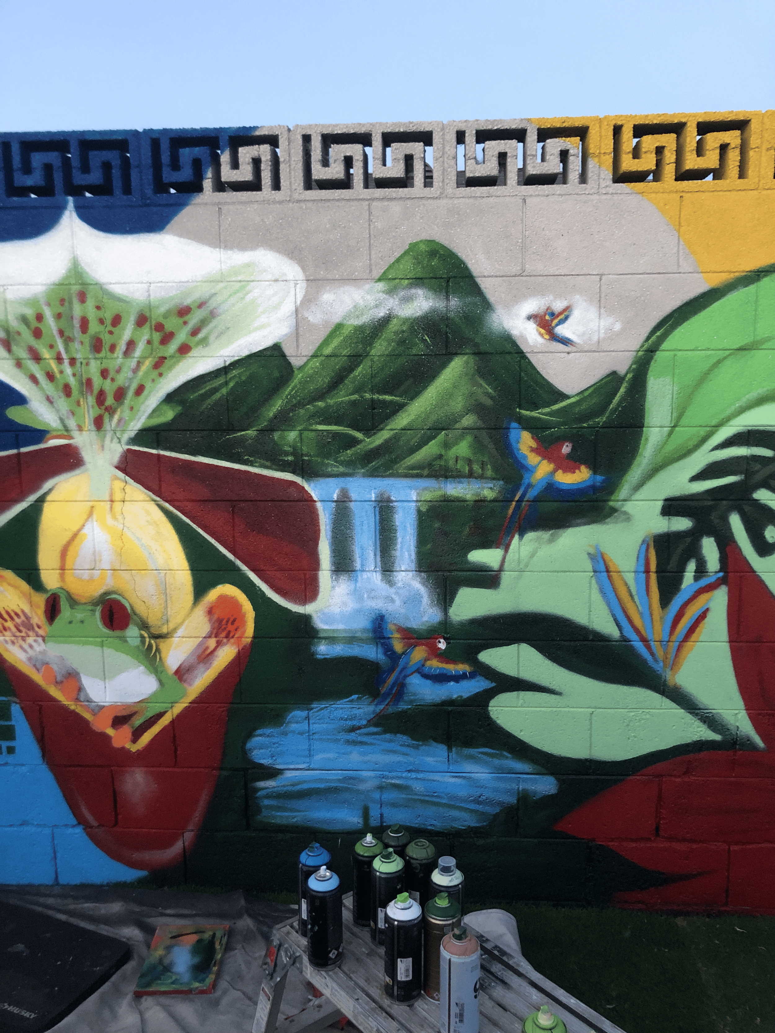 A colorful mural on a brick wall depicts green mountains, a waterfall, a flower with red and white petals, and several birds, with spray paint cans and art supplies in the foreground.