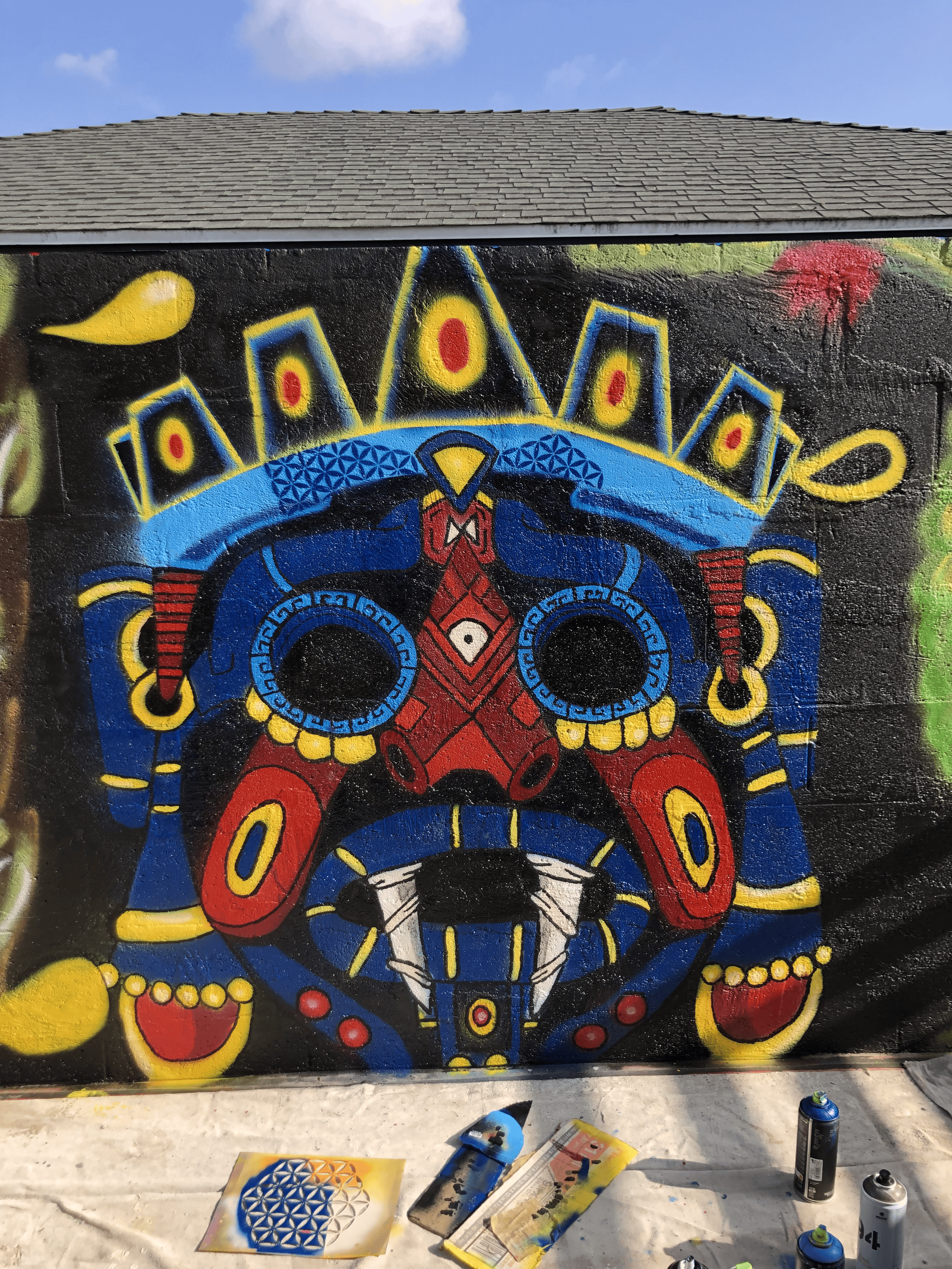 A colorful street art mural of a stylized, Talaloc Aztec Mask painted on a black wall with spray paint cans and art supplies on a white surface in foreground, and a blue sky with clouds above.