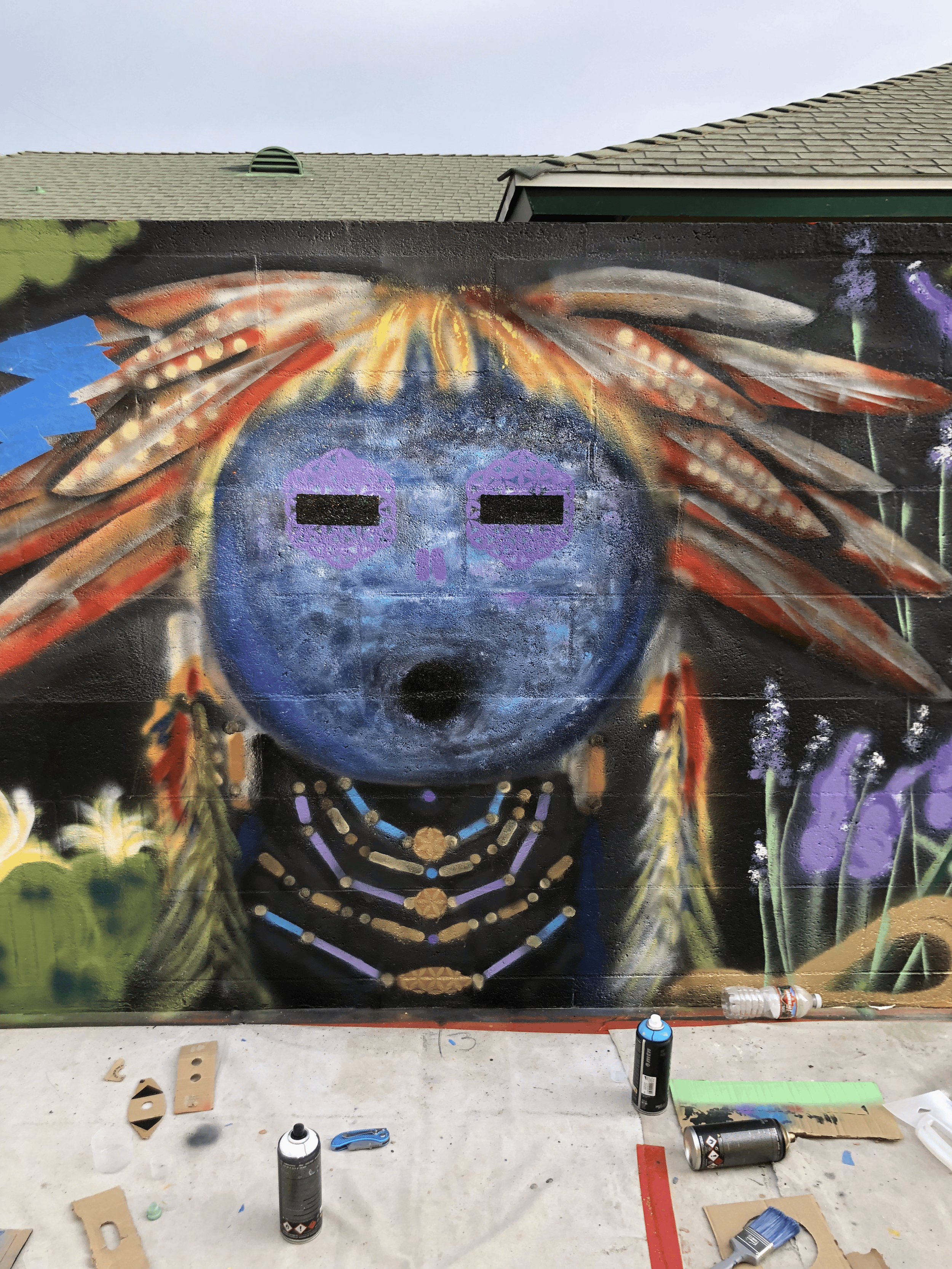 A colorful mural of a North American Native Tribe Mask, decorated with feathers and vibrant patterns, with art supplies like spray paint cans and brushes on the table below.