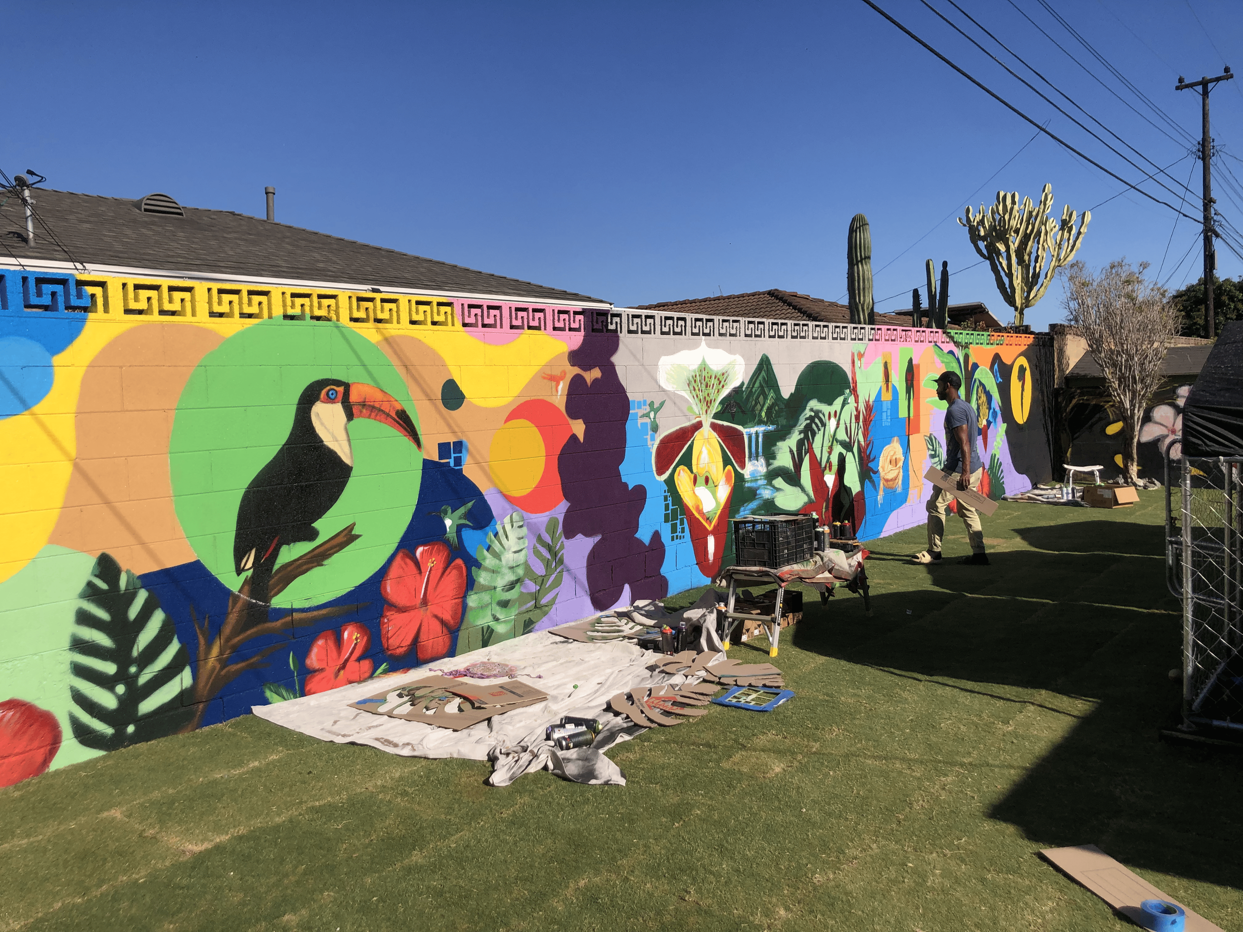 Colorful mural with tropical birds, flowers, and abstract patterns being painted on a wall outdoors, with art supplies on the ground and a man walking past.