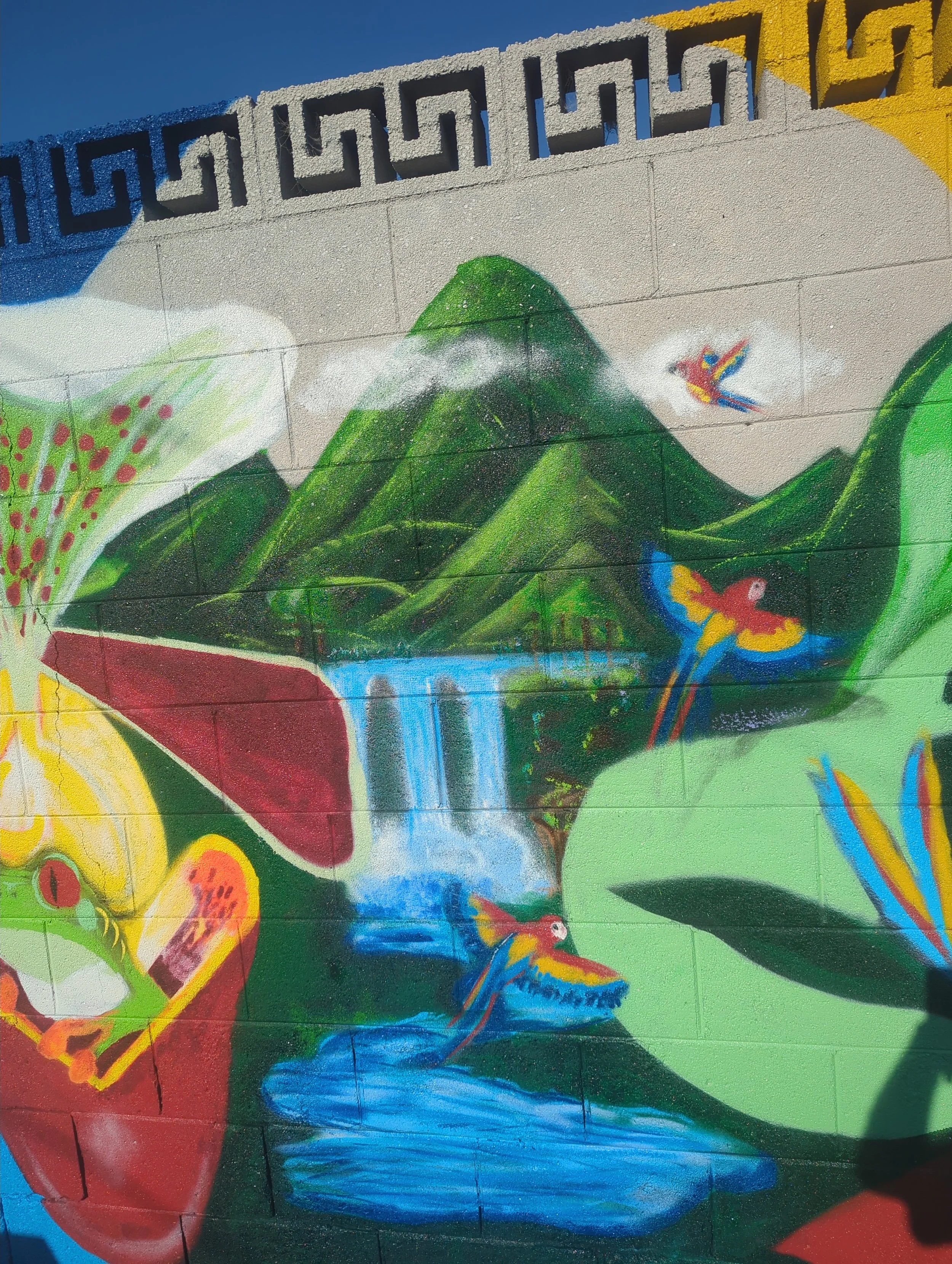 Colorful mural painting of a mountain range with a waterfall, surrounded by vibrant flora and fauna, including flying birds and butterflies, on a wall.