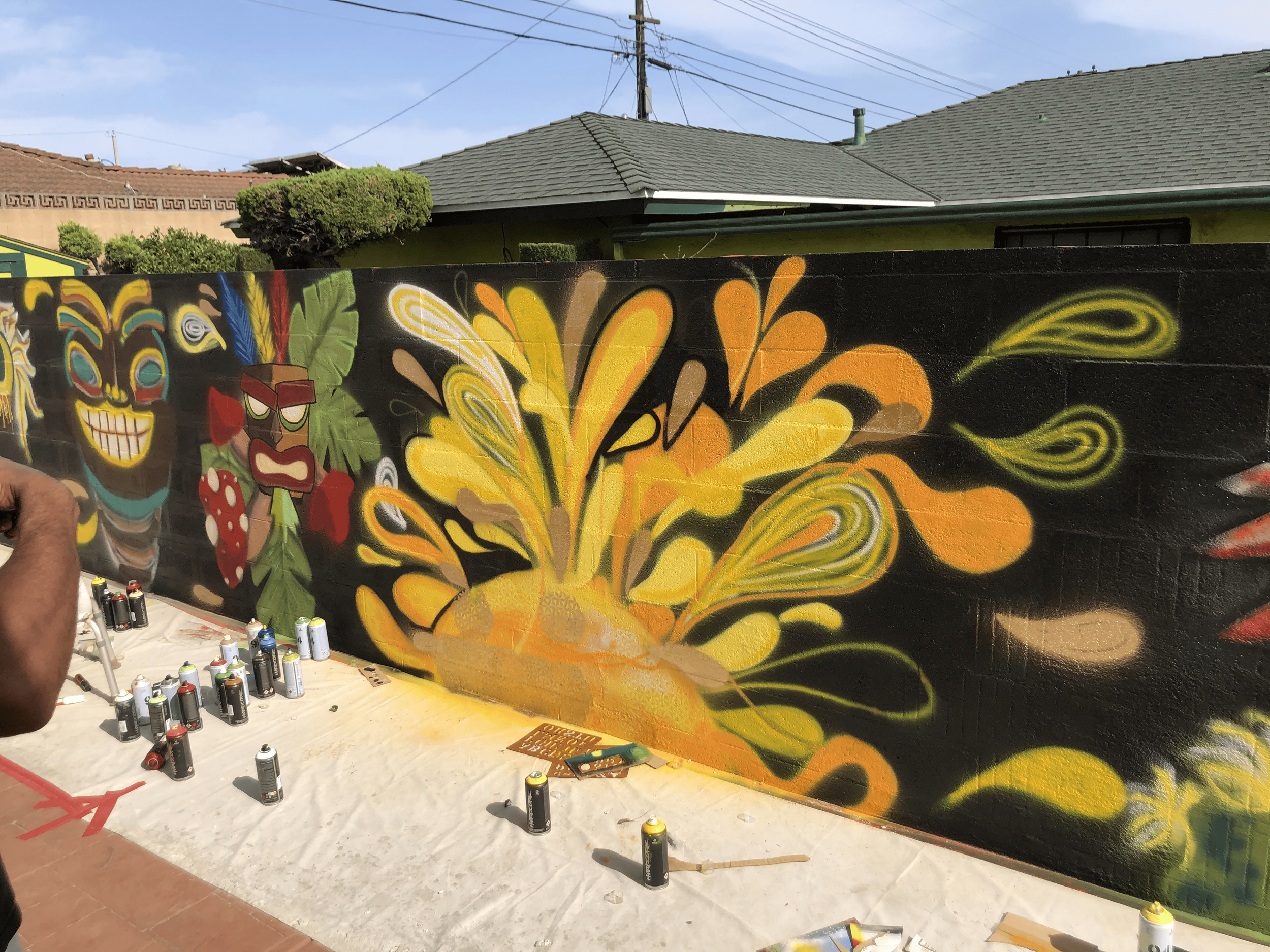 Colorful graffiti mural on a black wall featuring bright yellow, orange, and golden abstract sun with the flower of life patterning. Spray paint cans are scattered on the ground nearby.