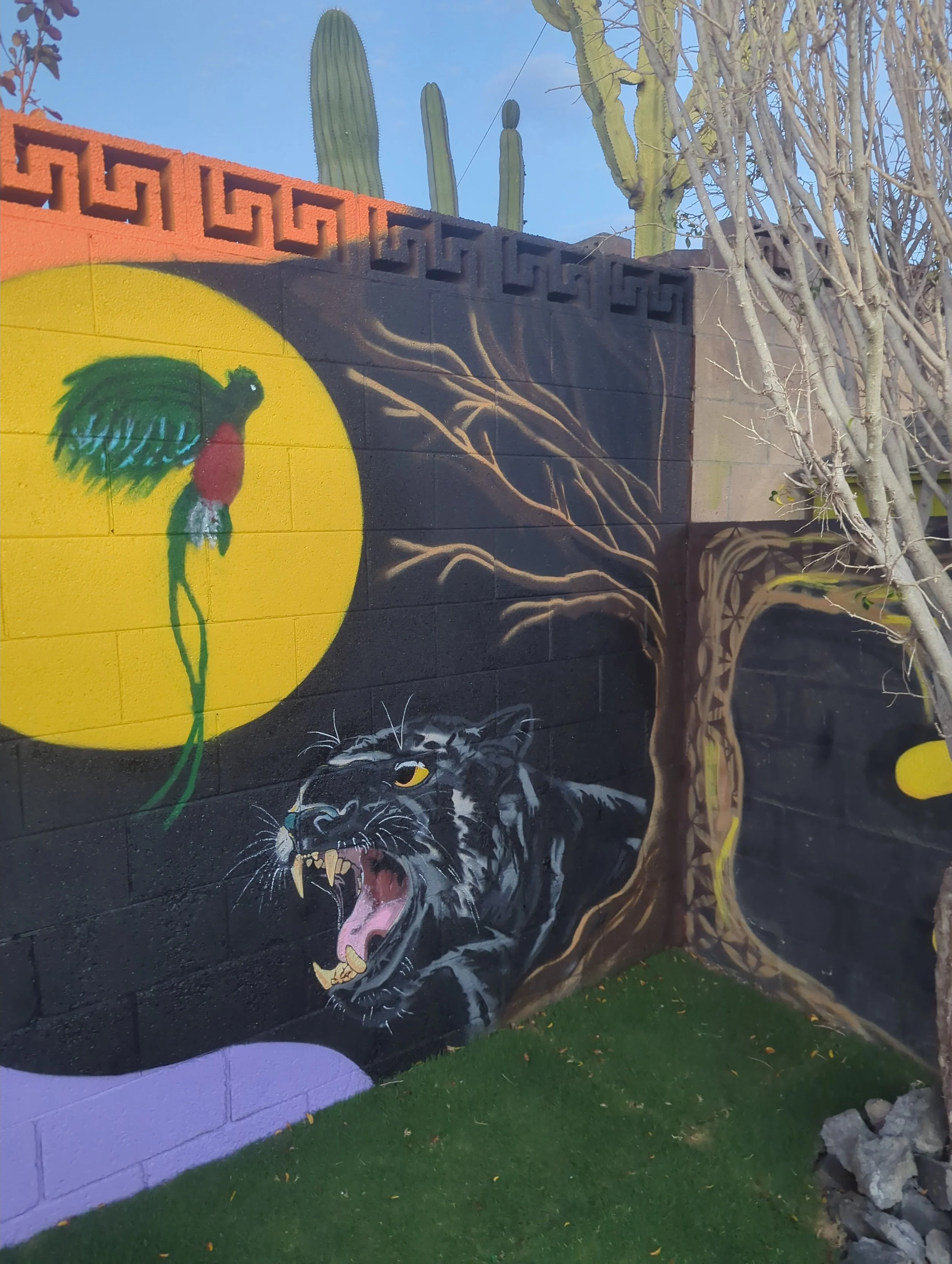 A colorful mural painted on a black brick wall features a black panther with yellow eyes and a roaring expression, a large green and red rooster with a yellow background, and a tree with roots extending downward, all set against the backdrop of cacti