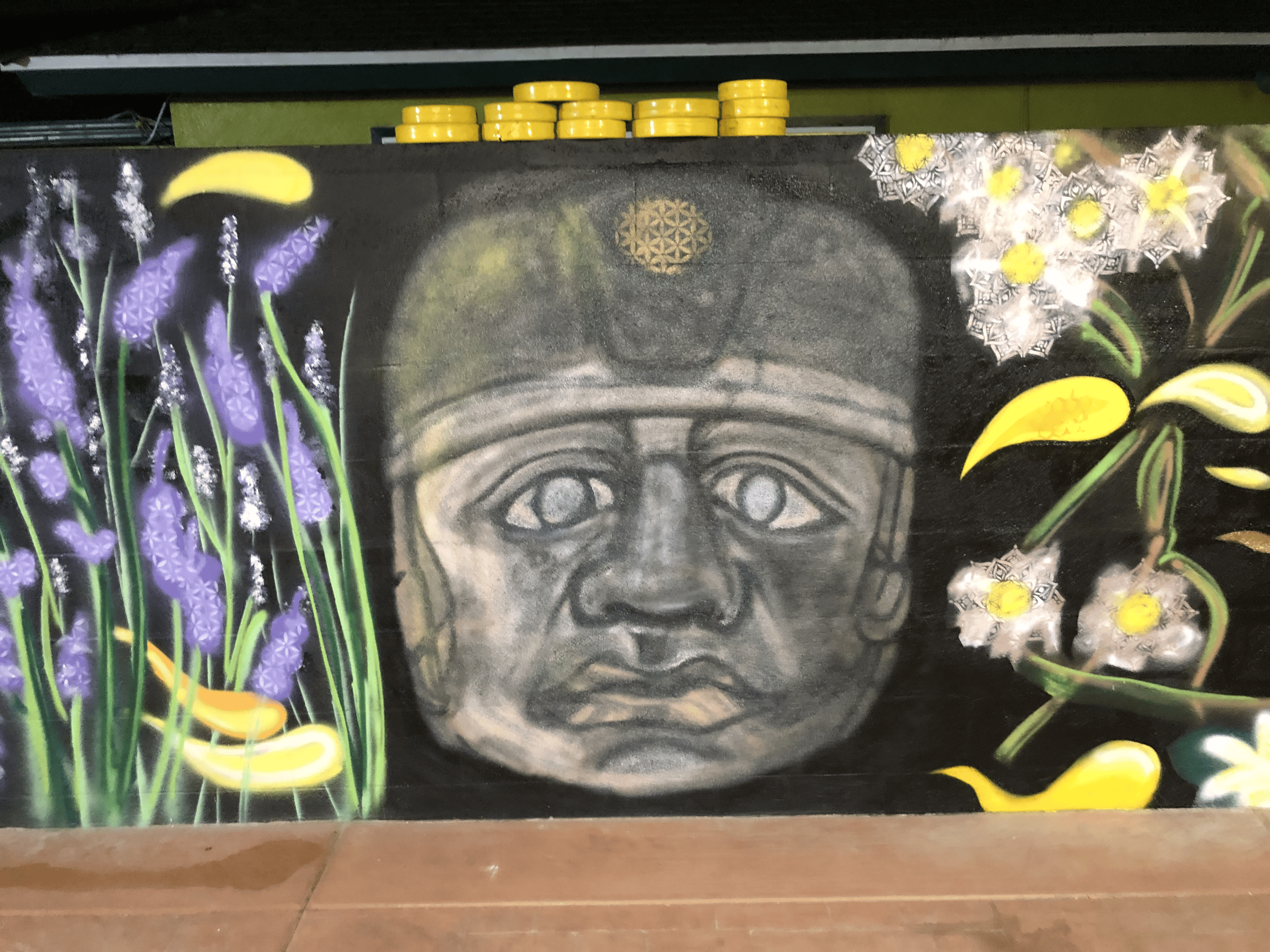 A mural featuring a stylized portrait of an Olmec Head, surrounded by purple lavender flowers and yellow lilies, and decorated with intricate black and white geometric patterns and yellow flowers.