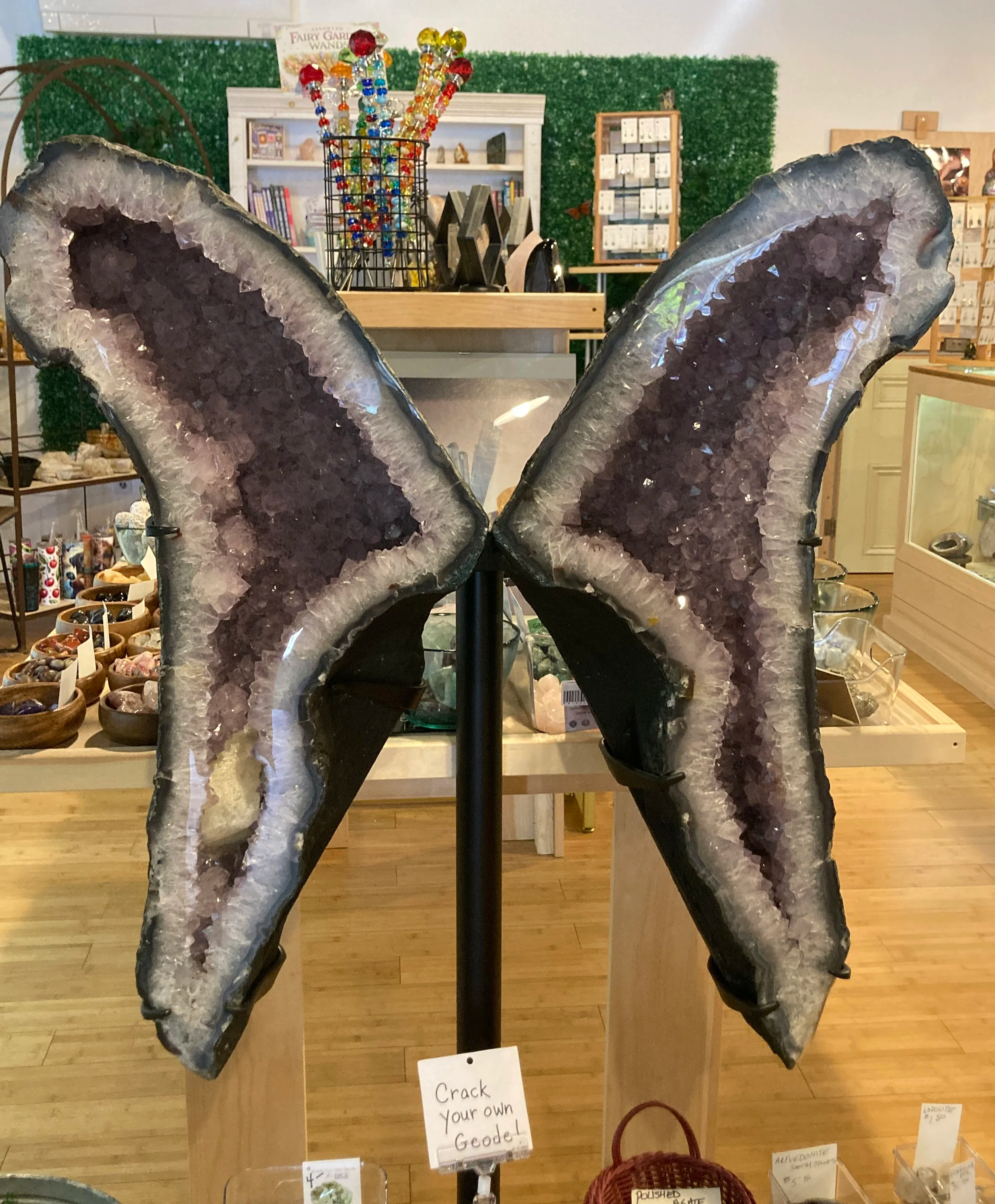 A display of large, purple, geode crystal slices with sparkling inner crystals in a store. The background includes shelves with jewelry, a green wall, and a sign that says "Crack your own Geode."