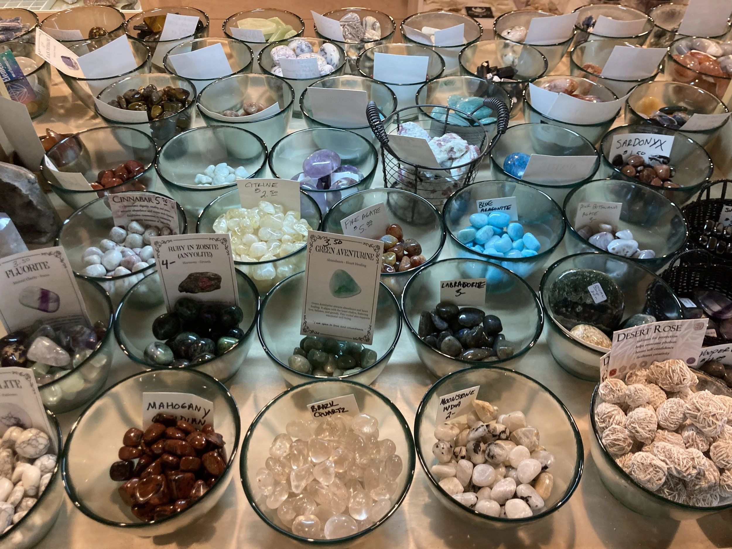 Multiple small glass bowls filled with colorful polished gemstones and crystals on a table, each with labels indicating their types and prices.