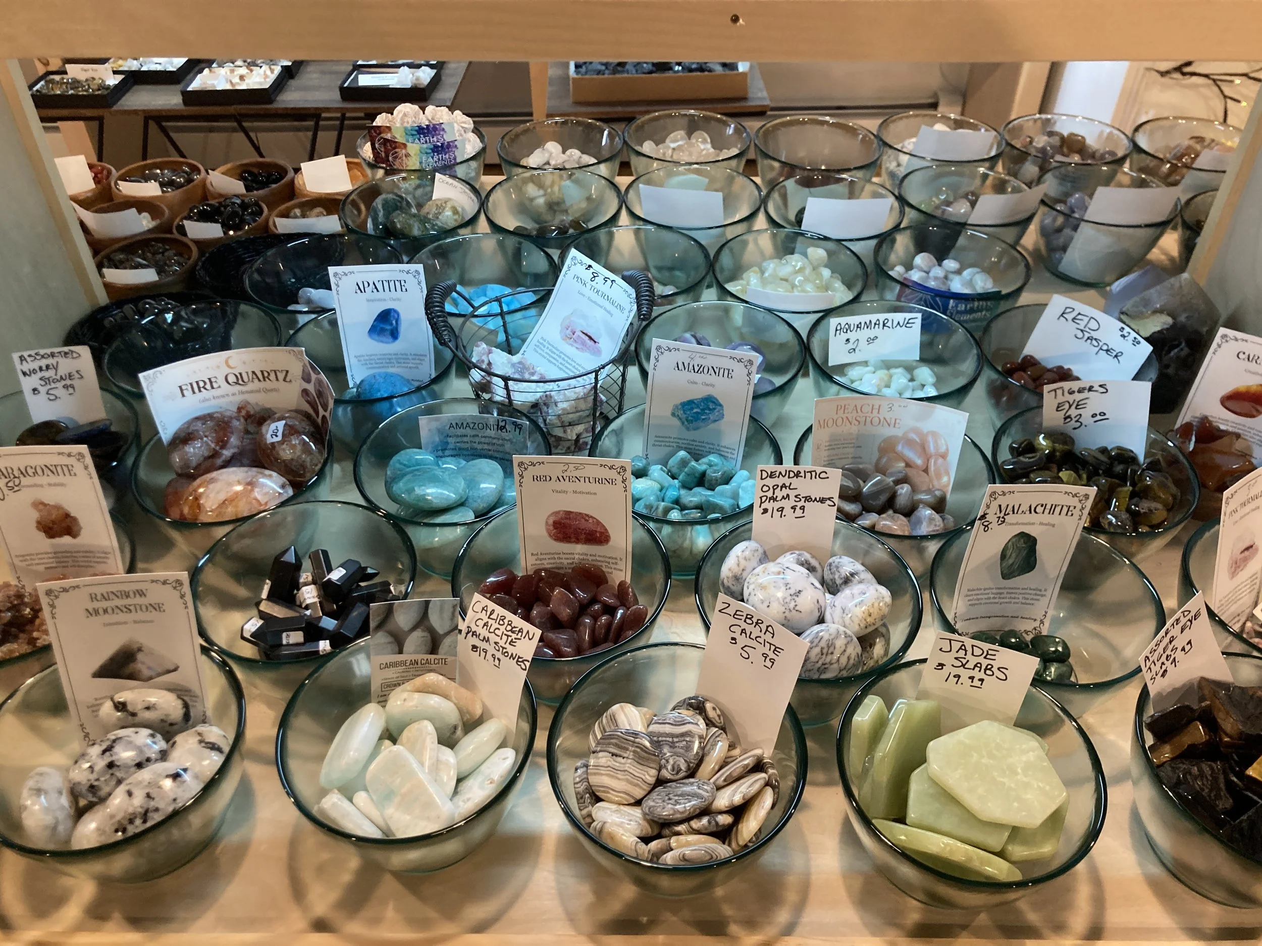 Display of various colorful polished stones and minerals in glass bowls at a store or exhibit, with labels and prices.
