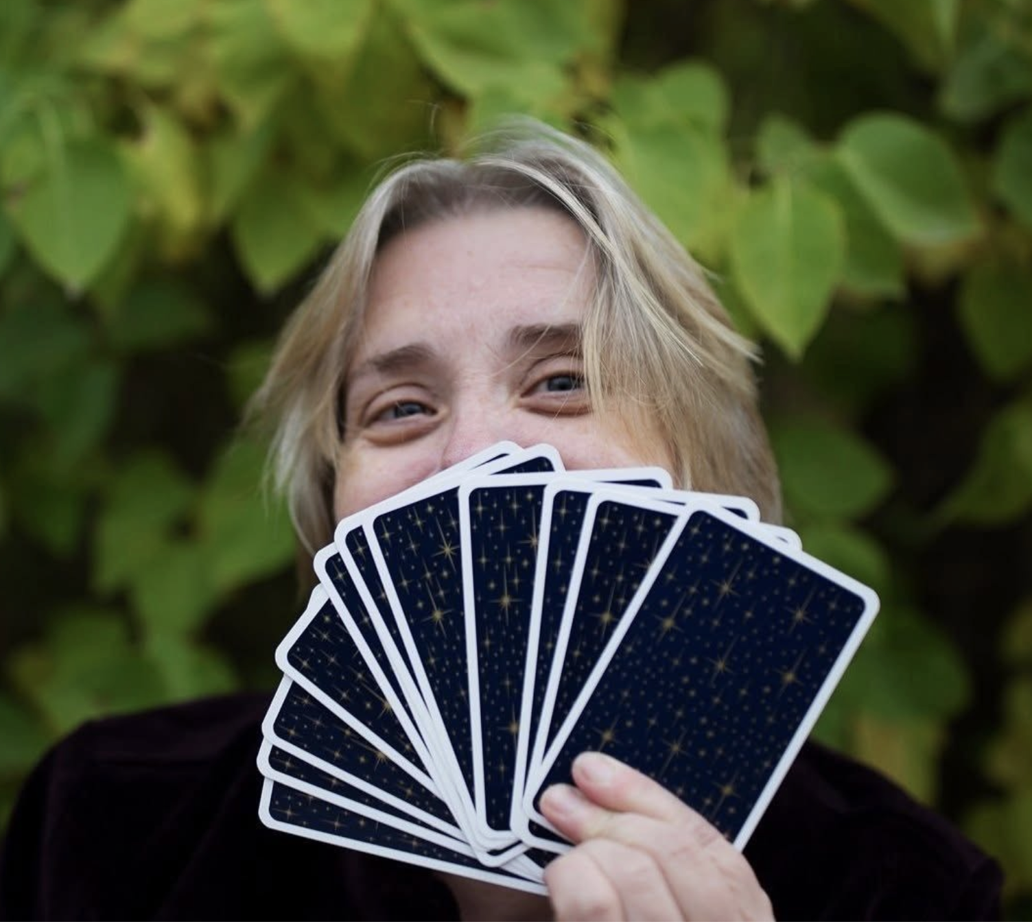 white woman with blong shaggy hair smiling and holding large black tarot cards in front of her face