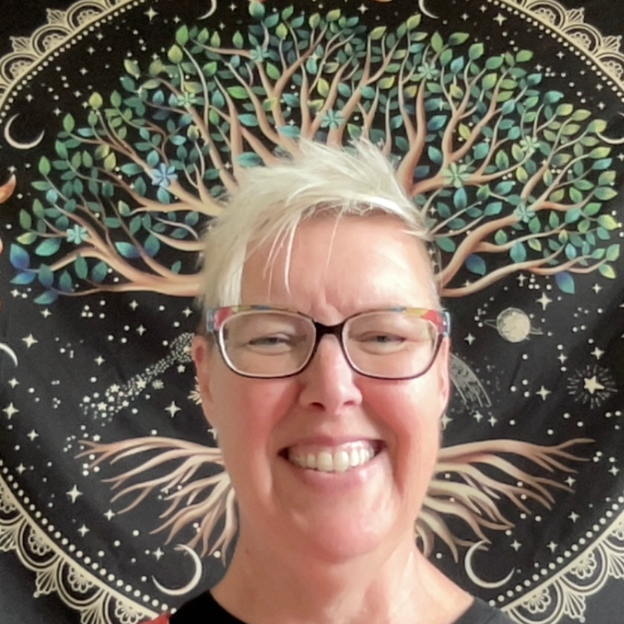 white woman with short blonde hair smiling in front of a tree of life wall hanging