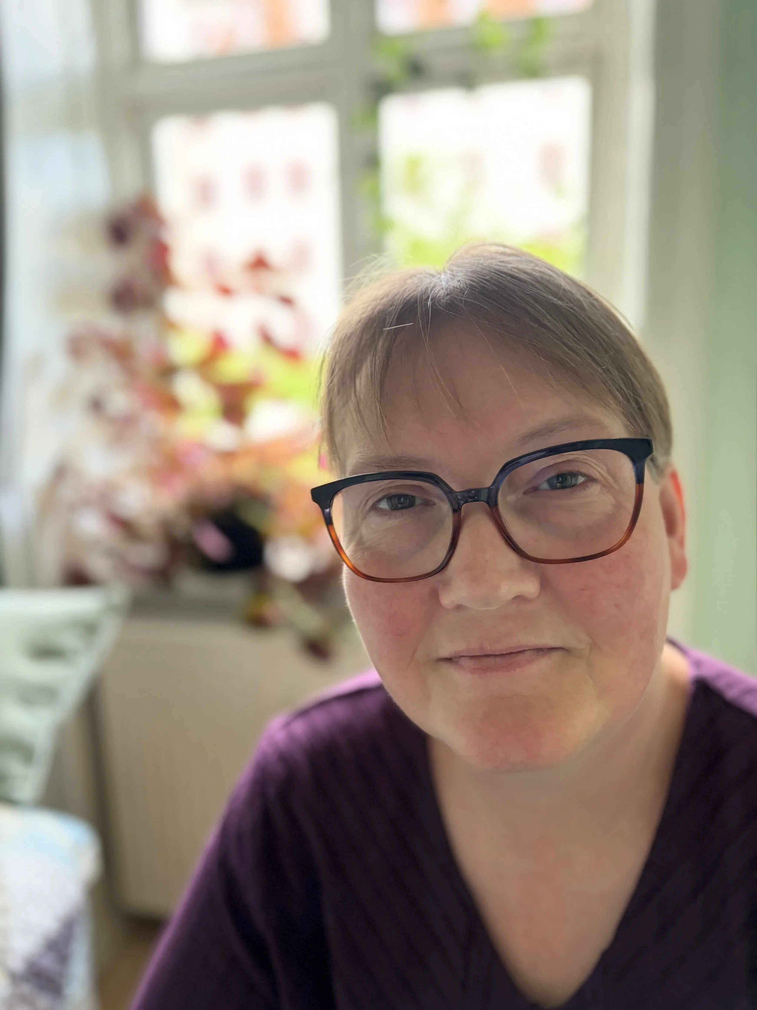 Billedet viser en kvinde med briller, der sidder i et rum med et vindue i baggrunden. I baggrunden er der en hylde med en dekoration eller planter.