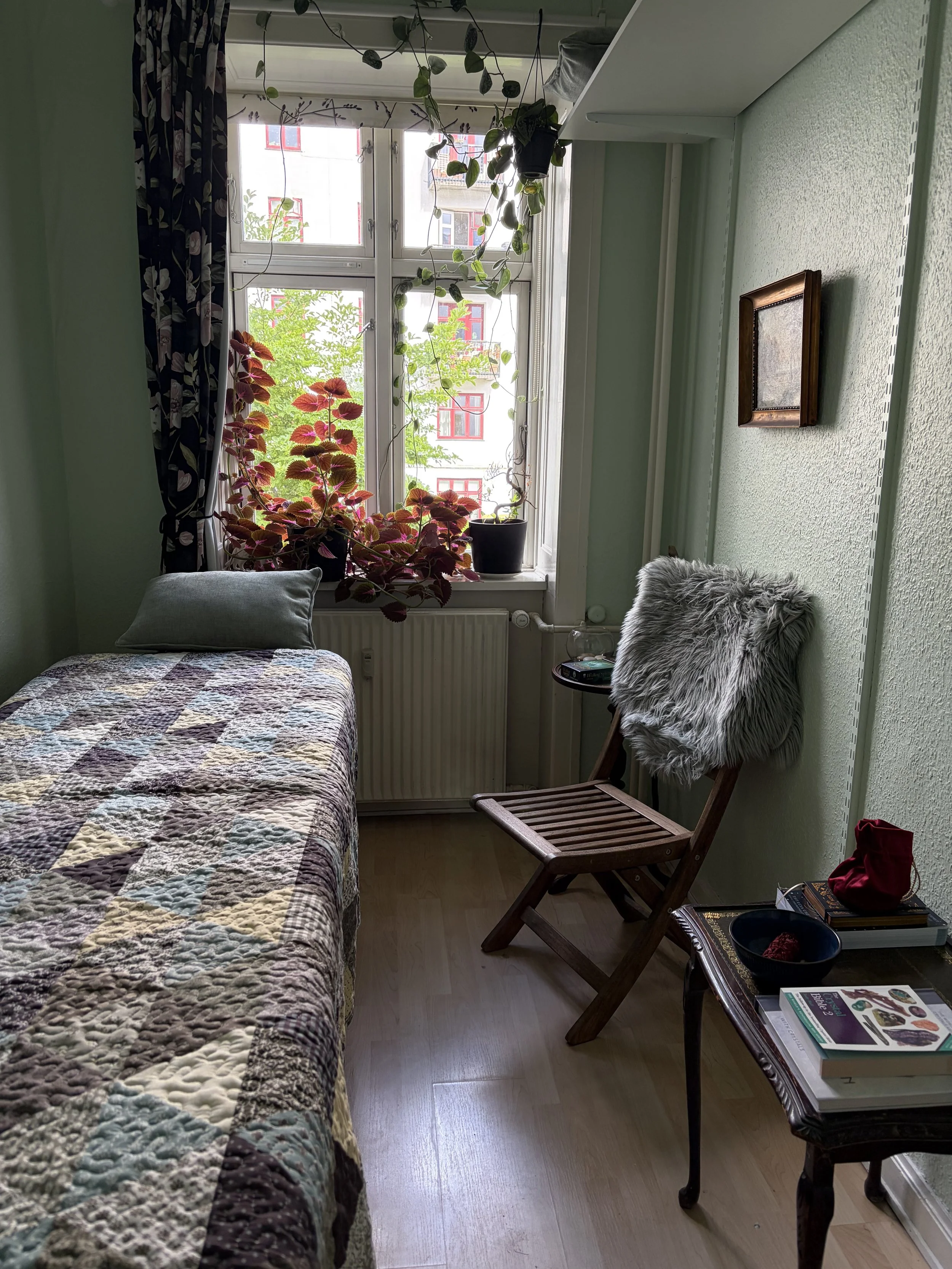 Billedet viser et lille værelse med en seng, der har et patchwork tæppe. Der er en pude på sengen. Ved siden af er der en vindueskarm med flere pottede planter. En træstol med en grå pels sætke sidder mod væggen. Der er en sidebord med bøger, en skål, og andre genstande. Vinduet har blomstrede mørke gardiner, og der er en bygning, der kan ses udenfor med røde vinduesrammer.