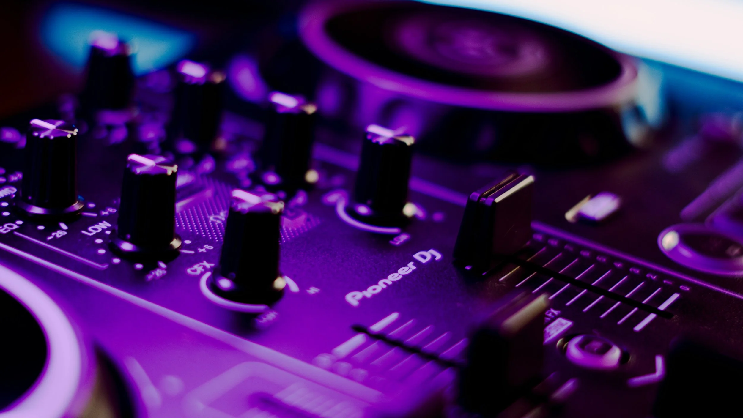 Close-up of a DJ mixer with purple lighting, showing control knobs, sliders, and turntable; Pioneer DJ branding visible.