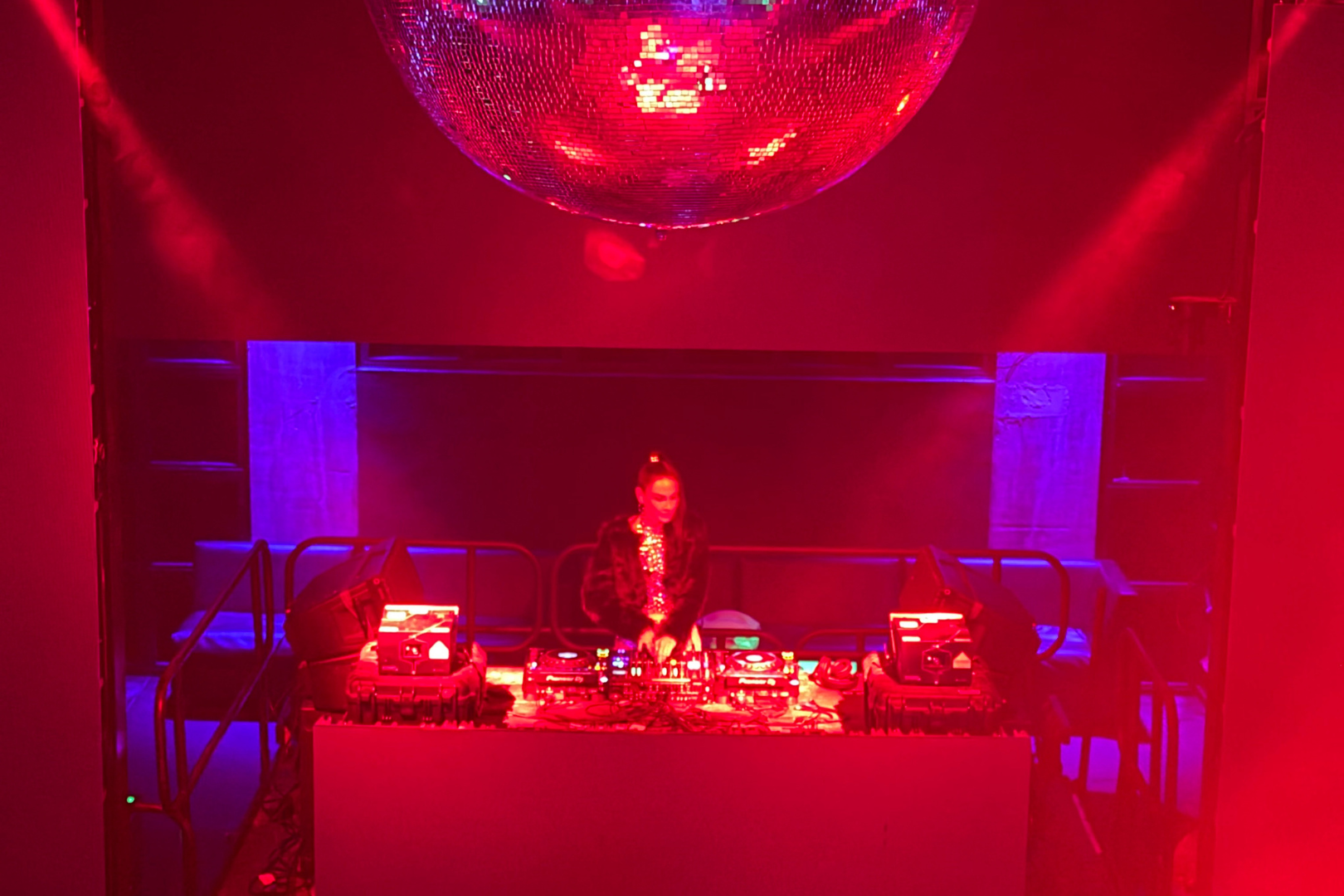 A DJ performs in a nightclub with red lighting, a large reflective disco ball hanging from the ceiling, and blue accent lights on the walls.