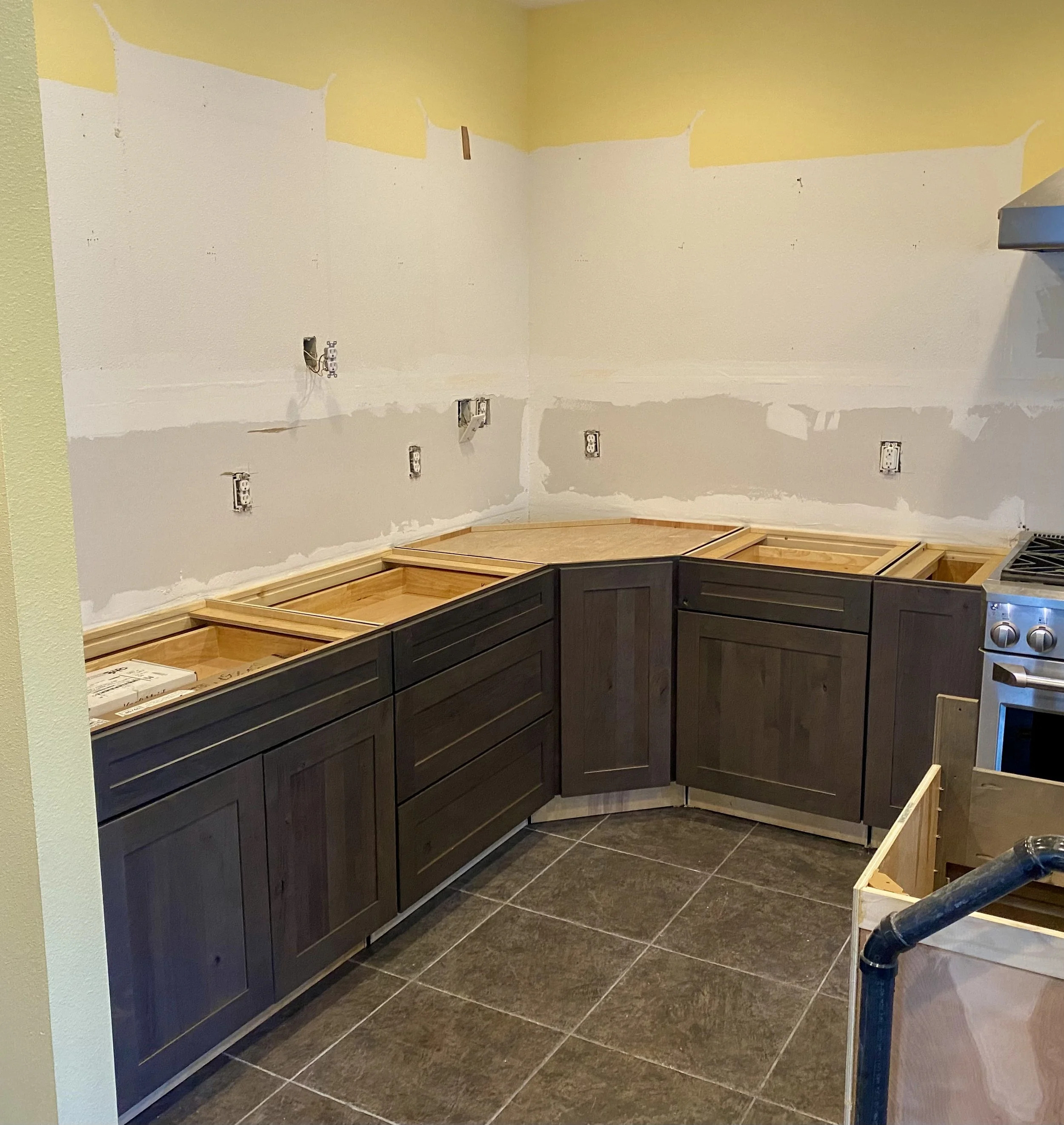 Kitchen with dark cabinets, unfinished walls, and electrical outlets. The upper walls are partially painted or have drywall work in progress, with some yellow paint near the ceiling. The floor is made of dark tiles, and some construction materials and appliances are visible in the corner.