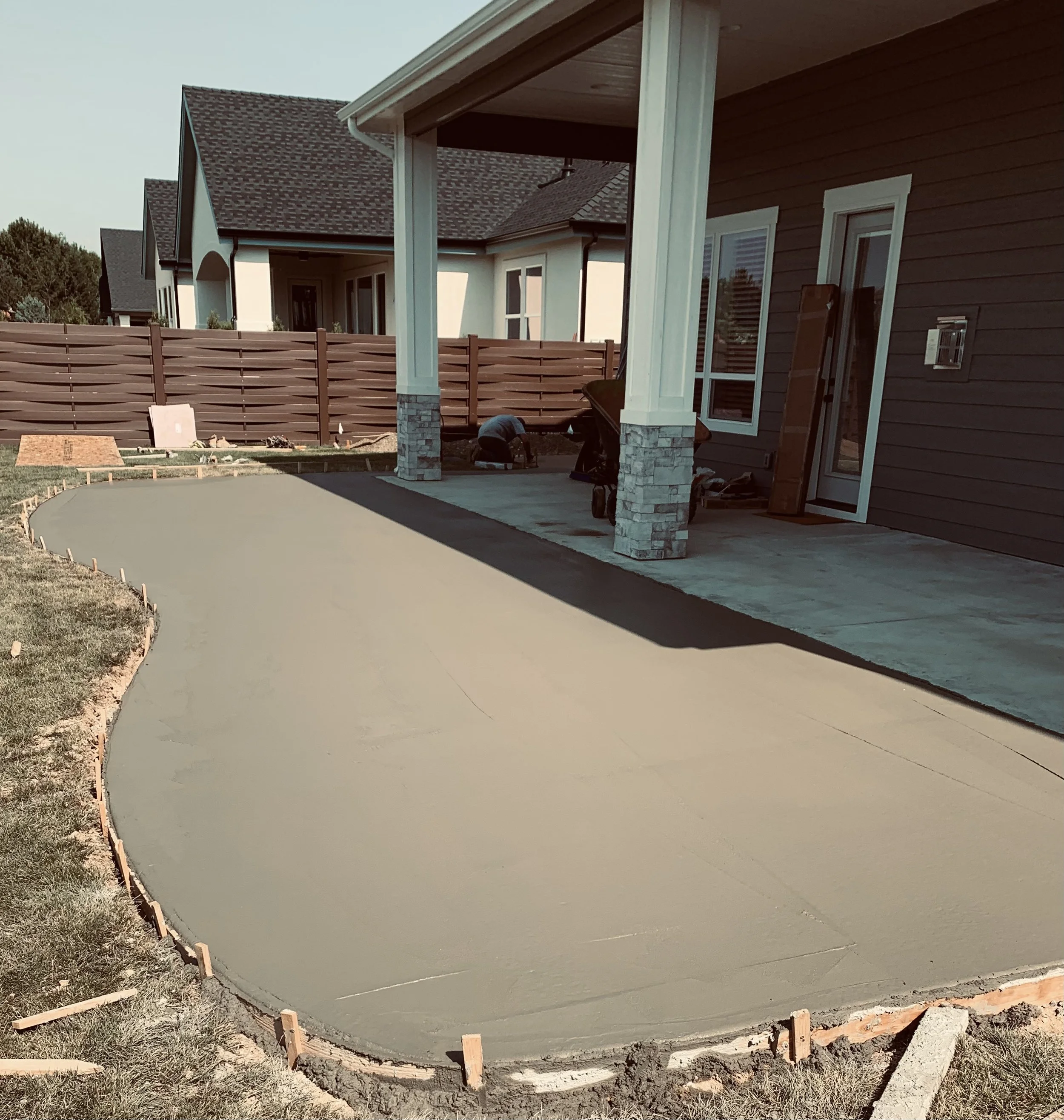 A newly poured concrete patio with curved edges in a backyard, surrounded by a wooden frame for finishing. There is a house with a porch and white trim on the right side, and new construction tools and materials are present on the ground.
