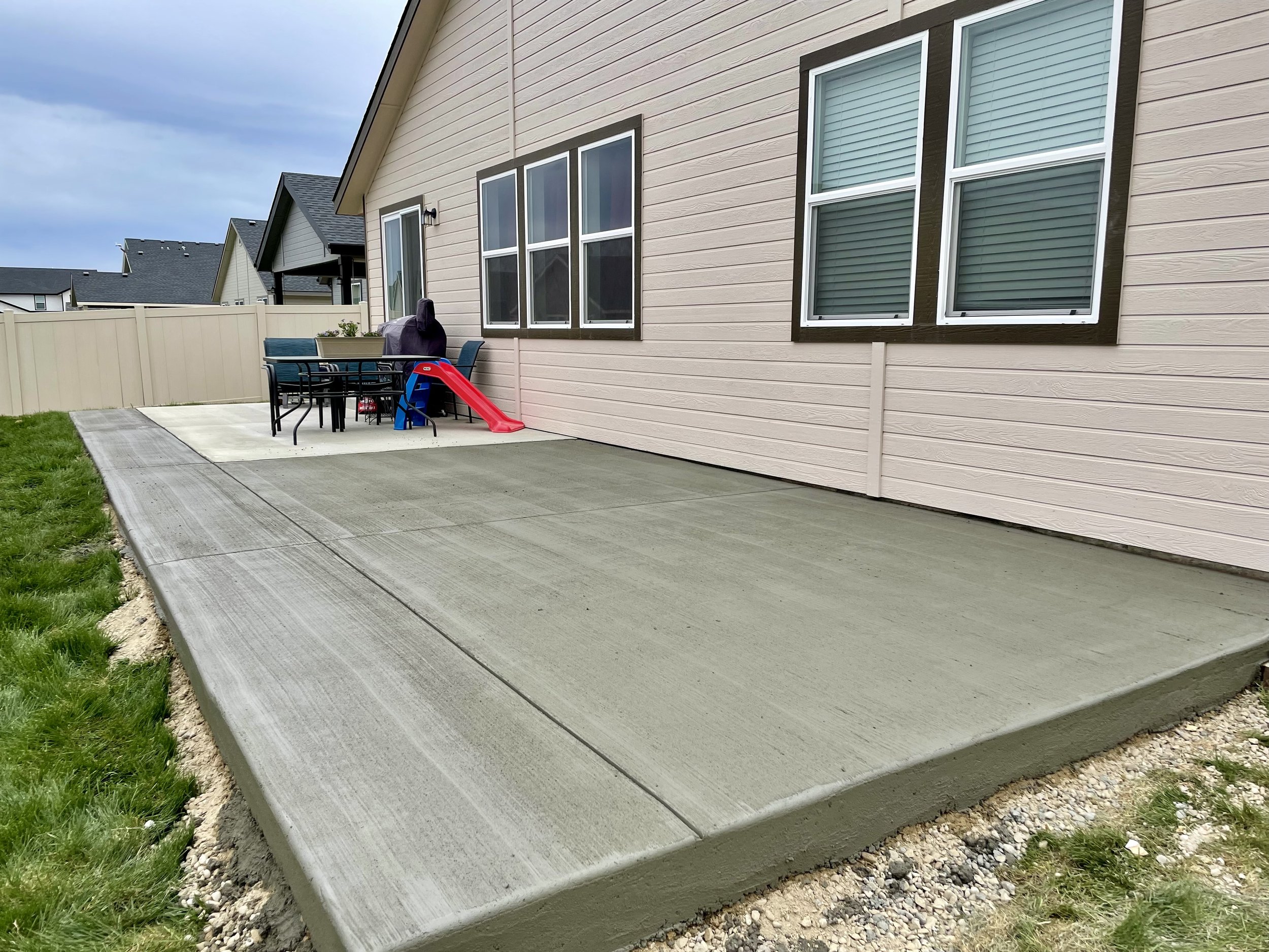 A concrete patio area outside a beige house with green grass border and outdoor furniture, including a table and chairs, and a small slide.