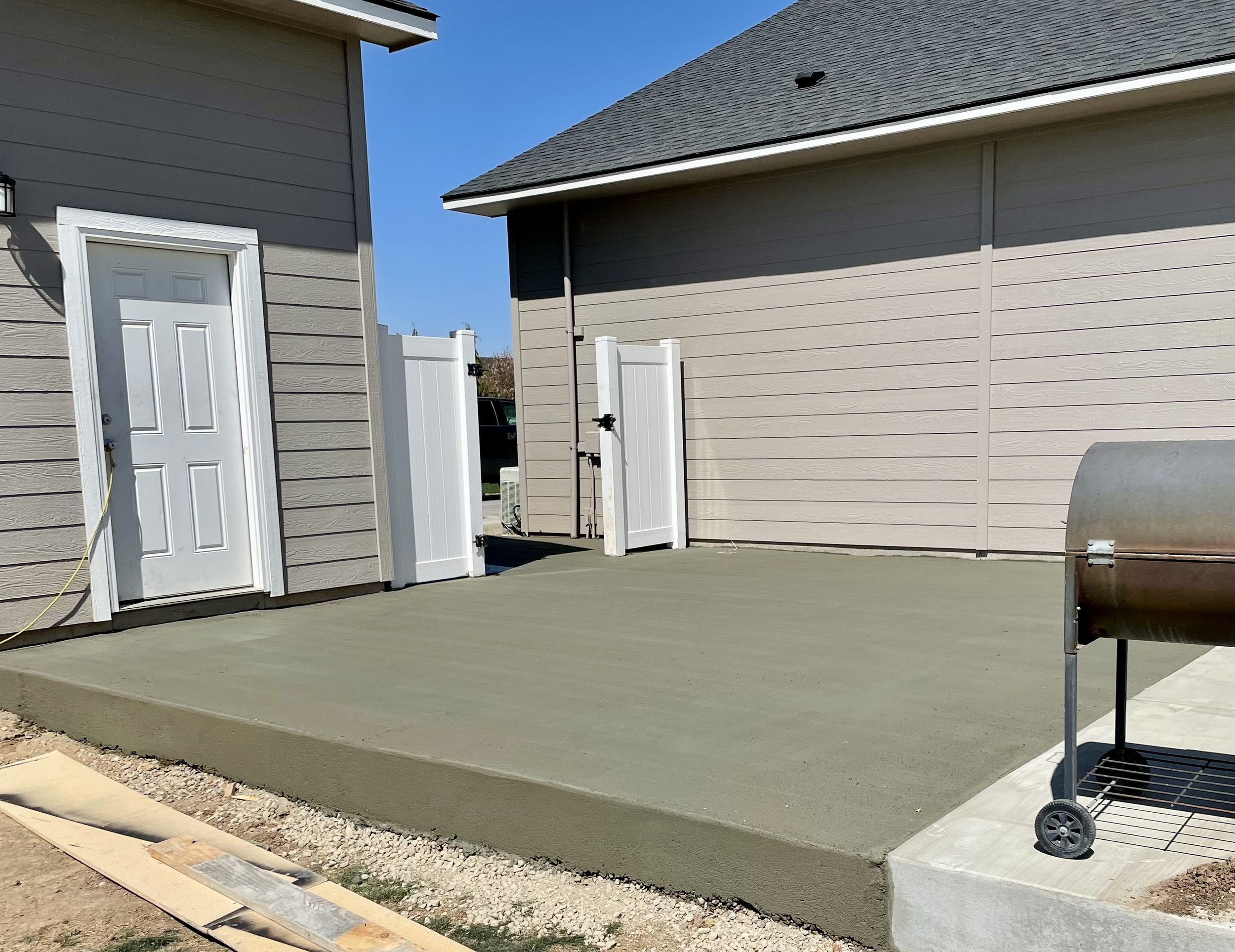 Concrete patio area between two house buildings with gray siding, a white door, a small black grill, and a white fence with a gate.