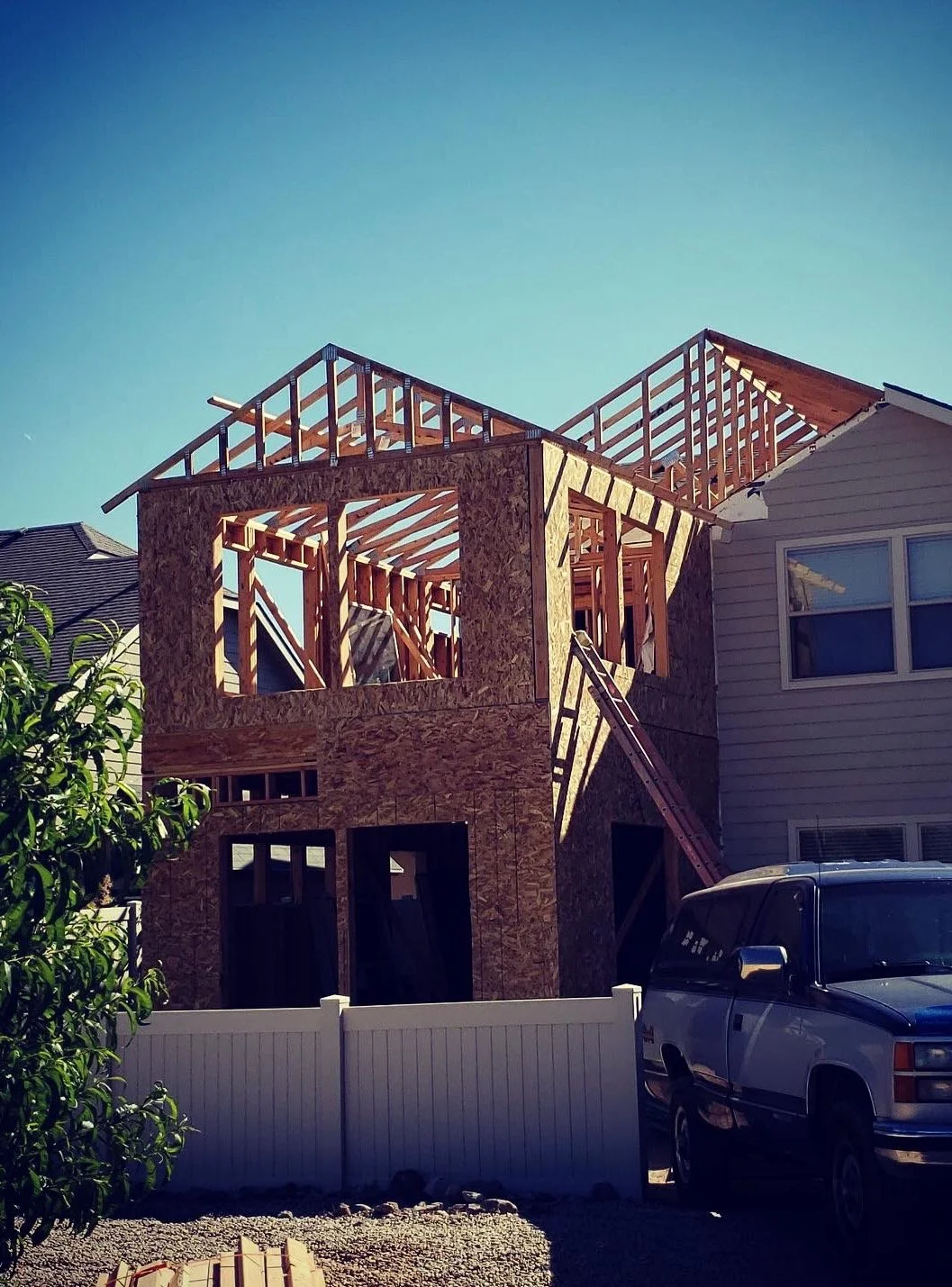 A house under construction with wooden framing, situated next to a finished house and a parked truck in the foreground.