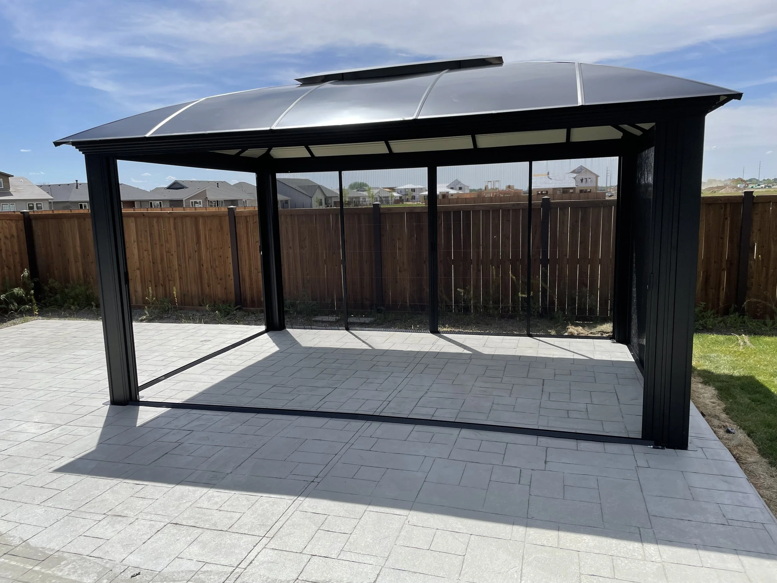 A backyard patio with a black metal and glass gazebo, gray brick pavers, a wooden privacy fence, and neighboring houses in the background under a partly cloudy sky.