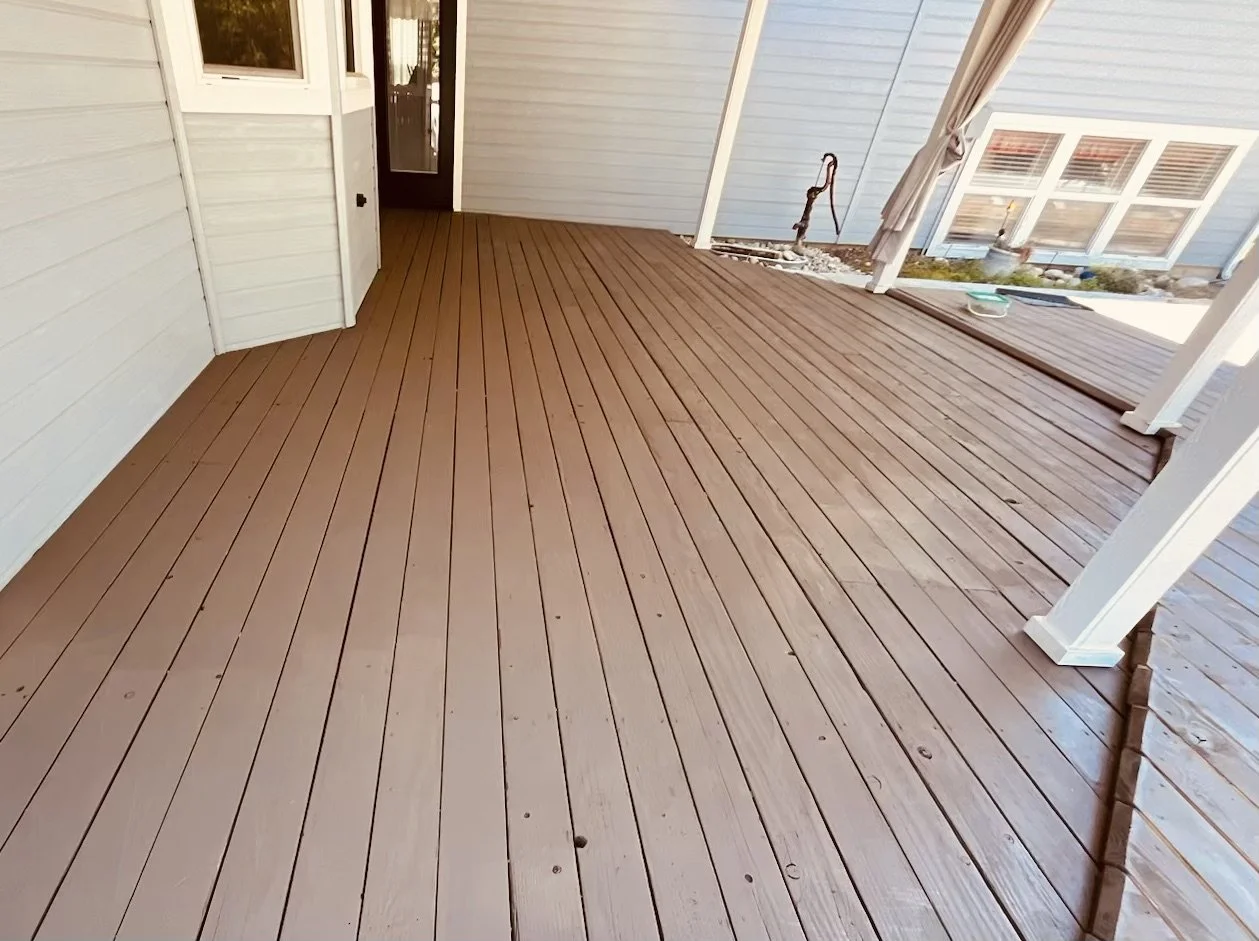 A newly built wooden deck attached to the house, with light blue siding, white trim, and a small patio area. There are a few plants, a garden hose, and a window visible in the background.