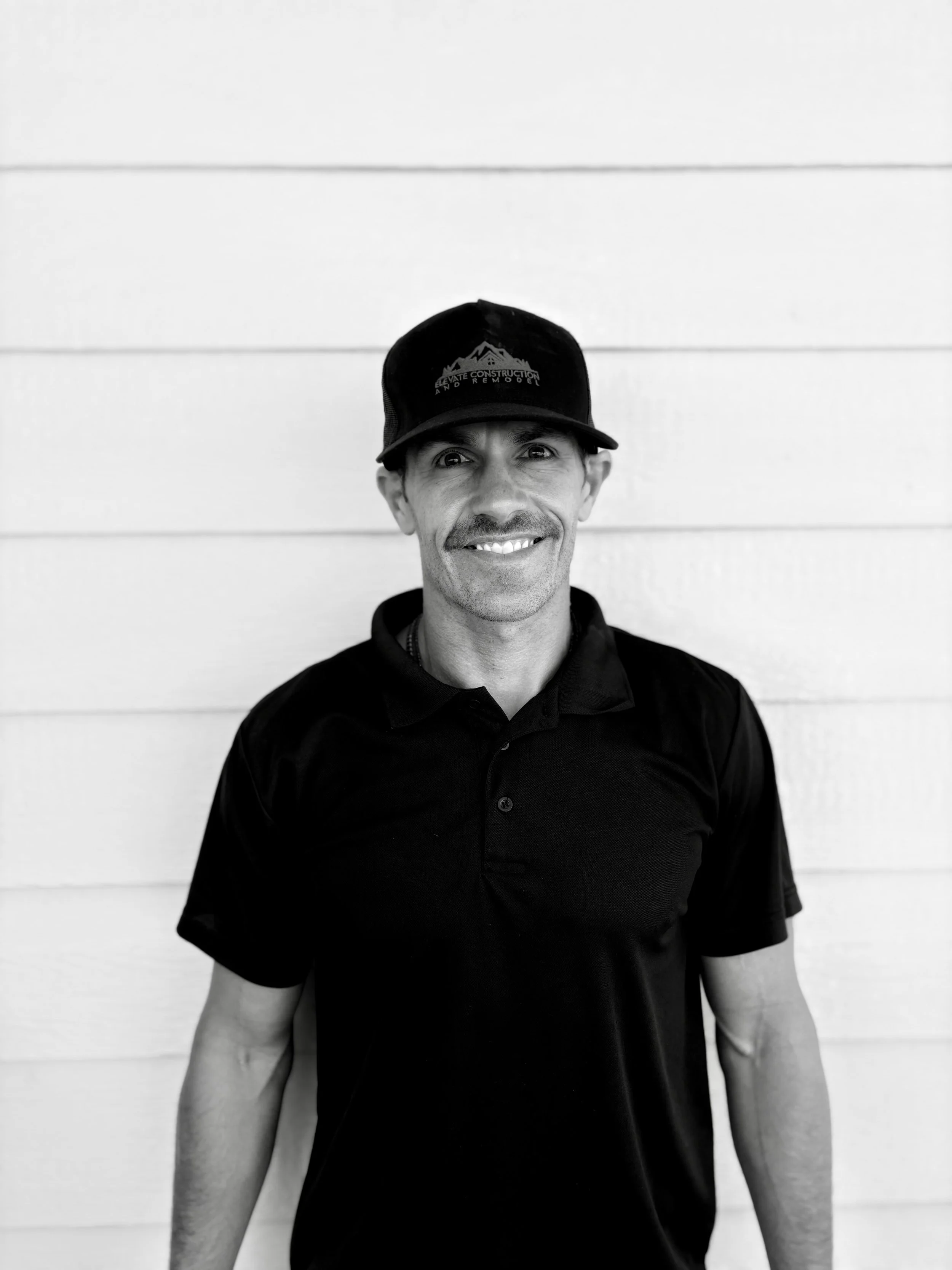 A smiling man wearing a black baseball cap and a black polo shirt standing in front of a white horizontal-striped wall.