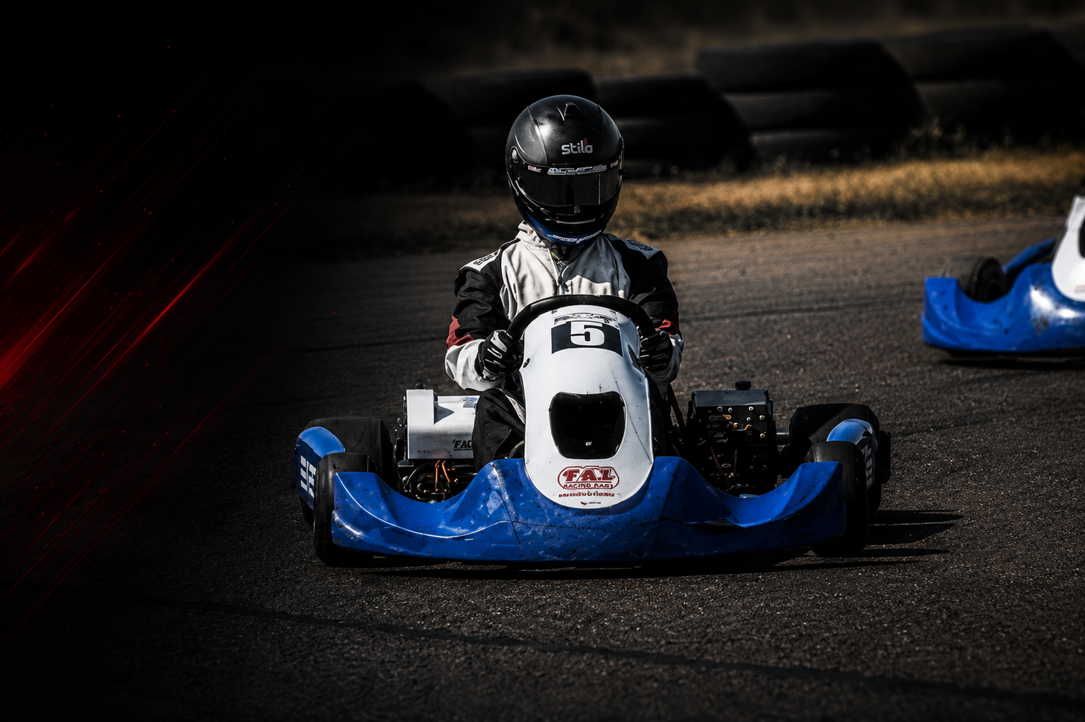 A person driving a go-kart on a race track, wearing a helmet and racing suit, with another blue go-kart visible in the background.