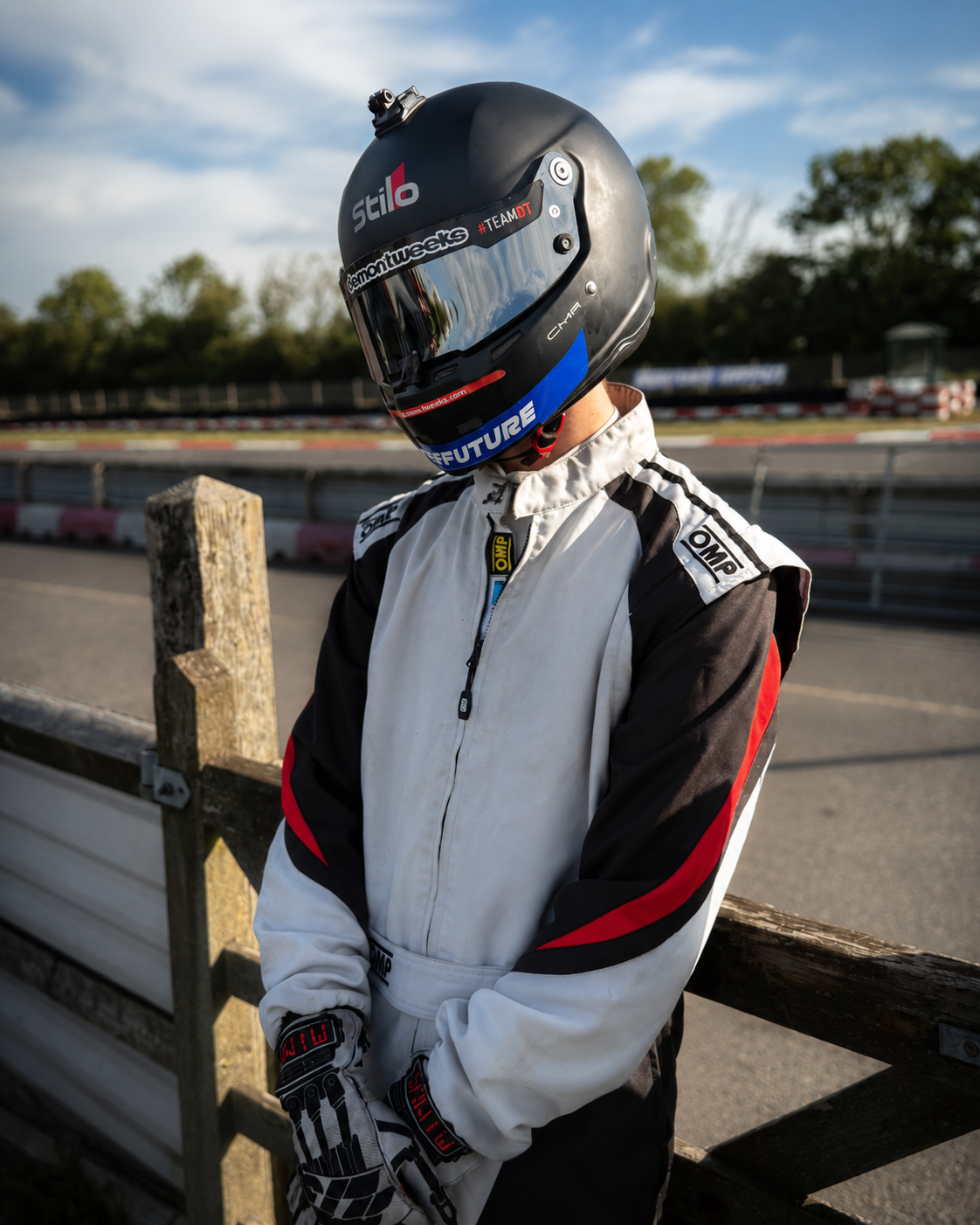 A race car driver standing near a racetrack, wearing a white racing suit with black and red accents and a black helmet with reflective visor.