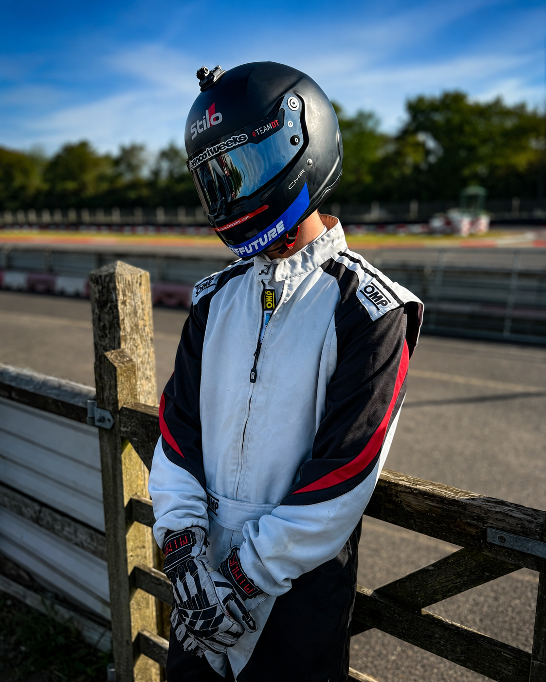 Person in racing suit and helmet standing near race track railing with trees and blue sky in background.