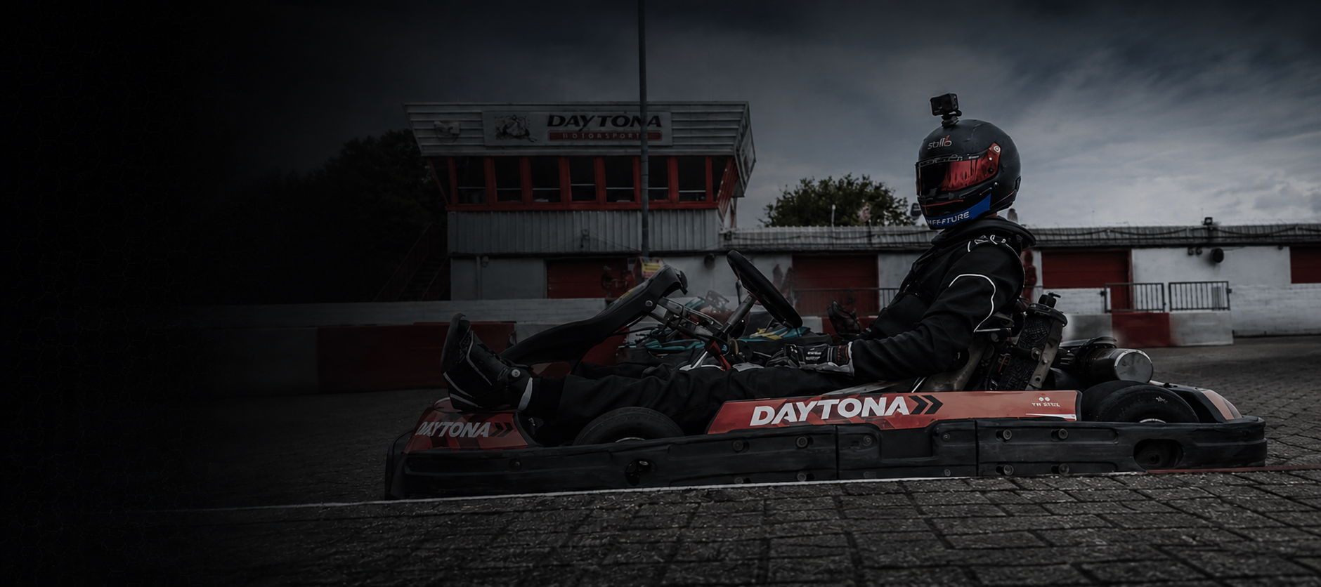 A person sitting on a go-kart with a helmet, at a race track, during dusk or dawn, with a building in the background labeled 'Daytona.'