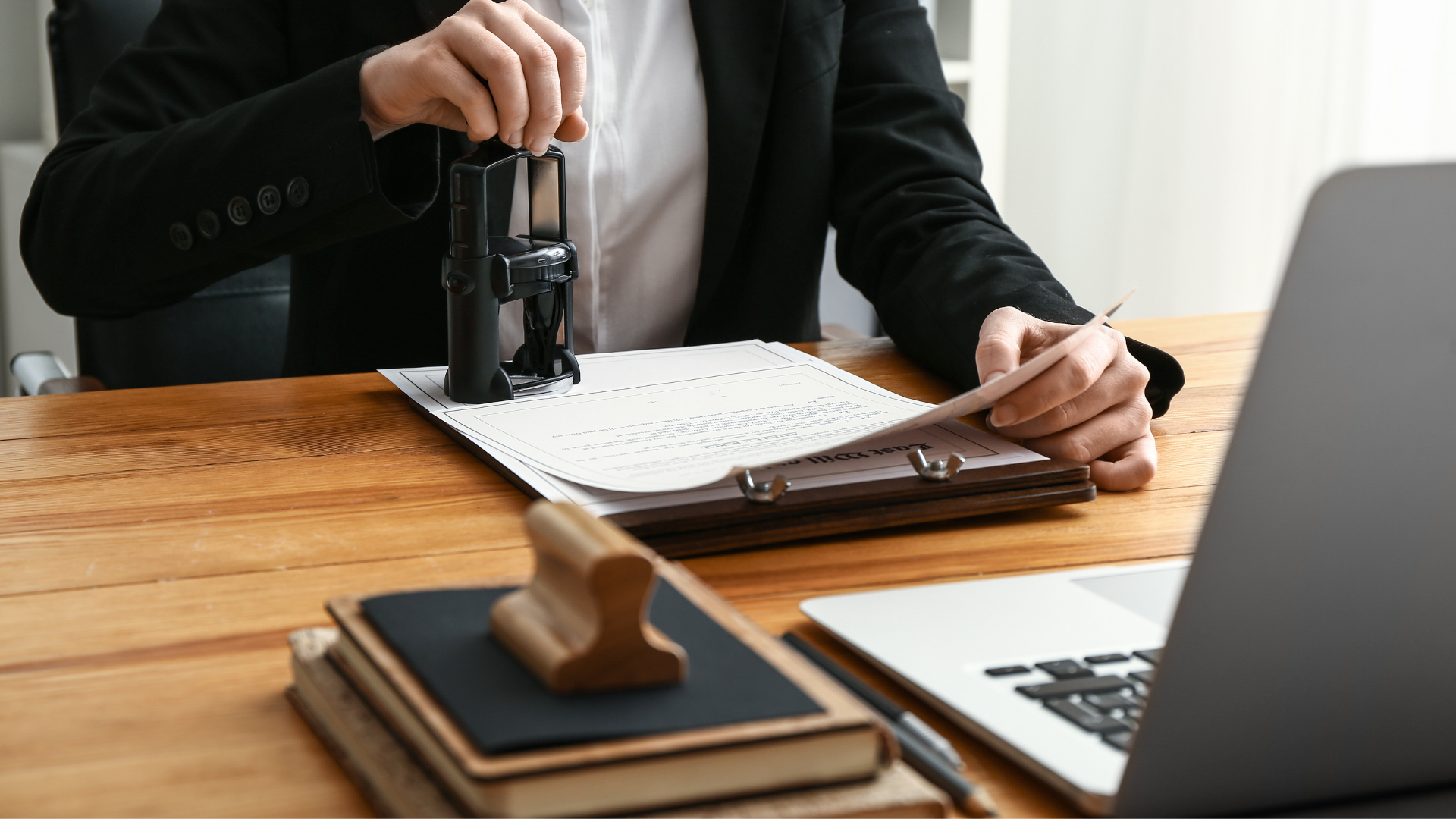Person in a suit reviewing documents at a wooden desk, with a stamp and a laptop nearby.