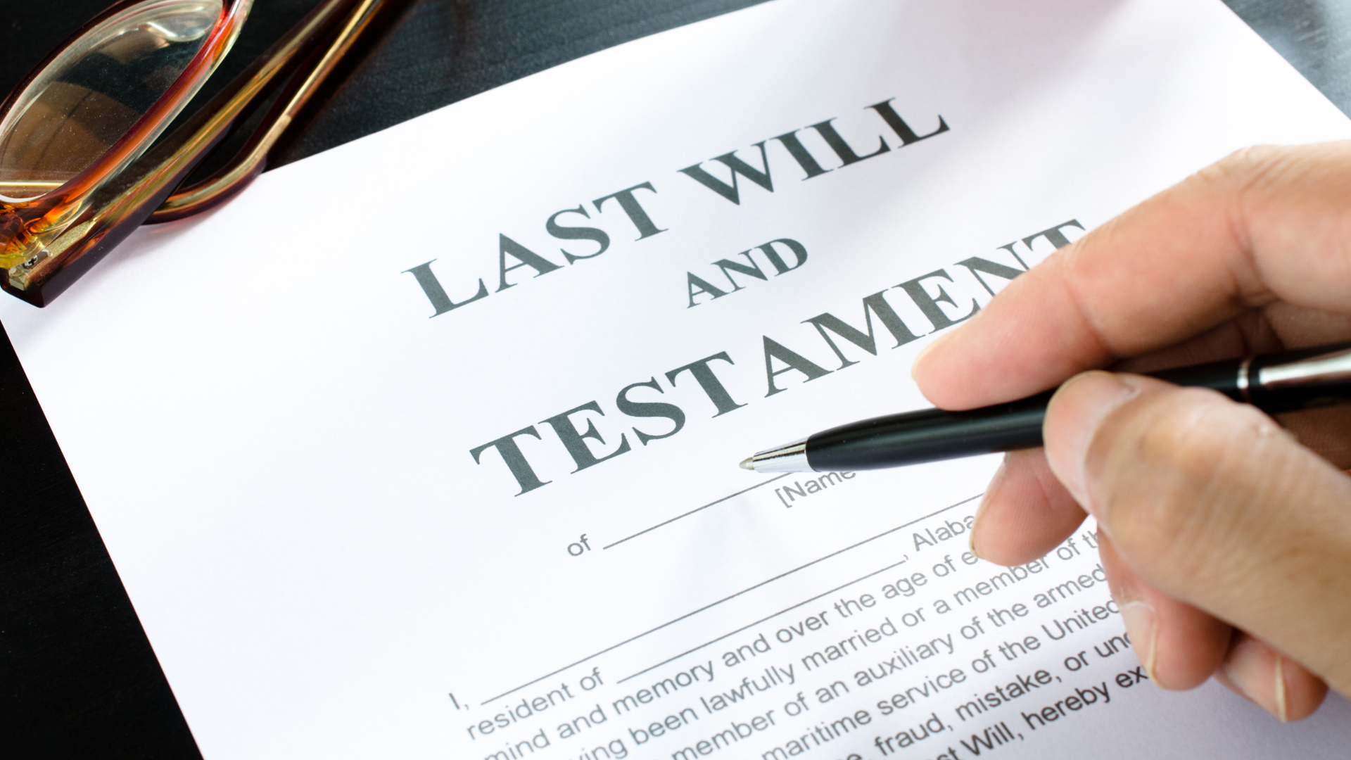 A close-up image of a legal will document titled "Last Will and Testament" with a person signing it using a black pen. A pair of tortoiseshell glasses rests on the top left corner of the paper.