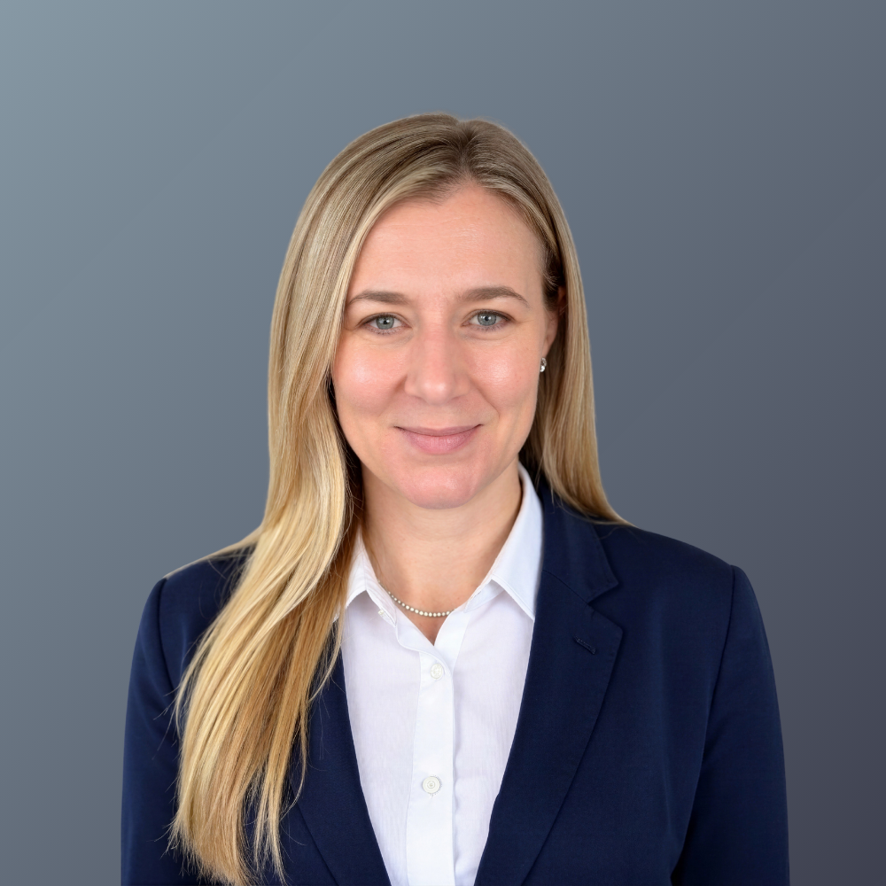 Professional woman with long blonde hair wearing a navy blazer and white shirt, smiling at the camera against a gray background.