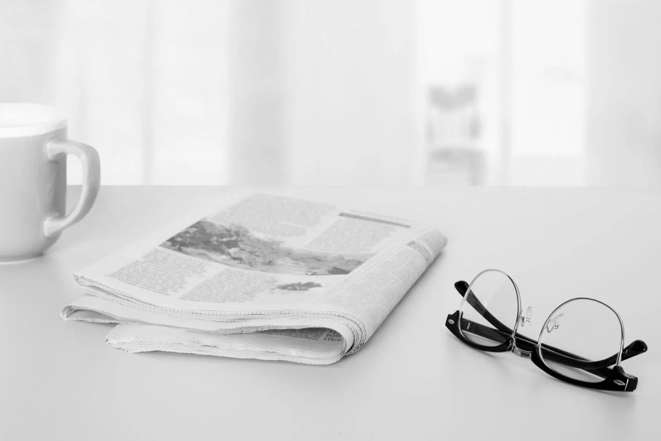 Une tasse blanche, un journal plié, et une paire de lunettes rectangulaires à monture noire sur une surface blanche, avec un fond flou.