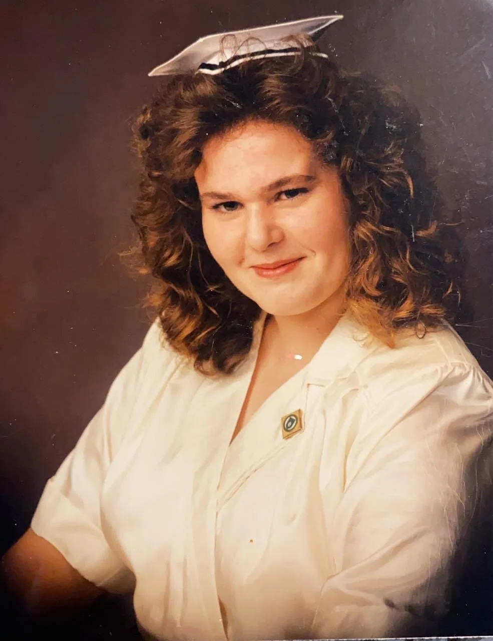 A woman with curly brown hair and fair skin, wearing a beige shirt and a small pin, is smiling at the camera against a dark background. She has a piece of paper or card on her head.