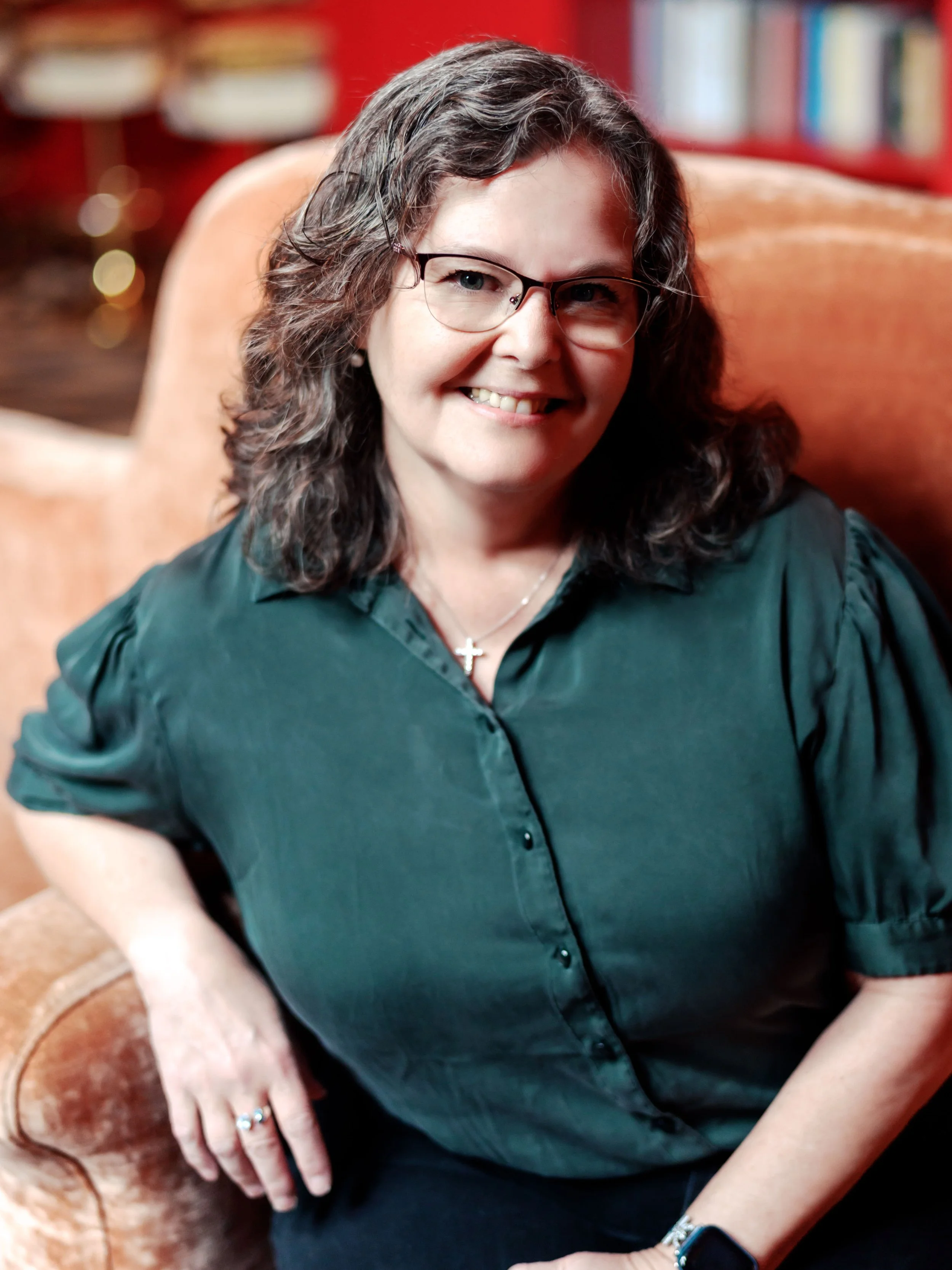 Close-up of a woman with curly gray hair, glasses, wearing a dark green blouse, smiling, sitting on a brown leather chair with a background of red and beige tones.