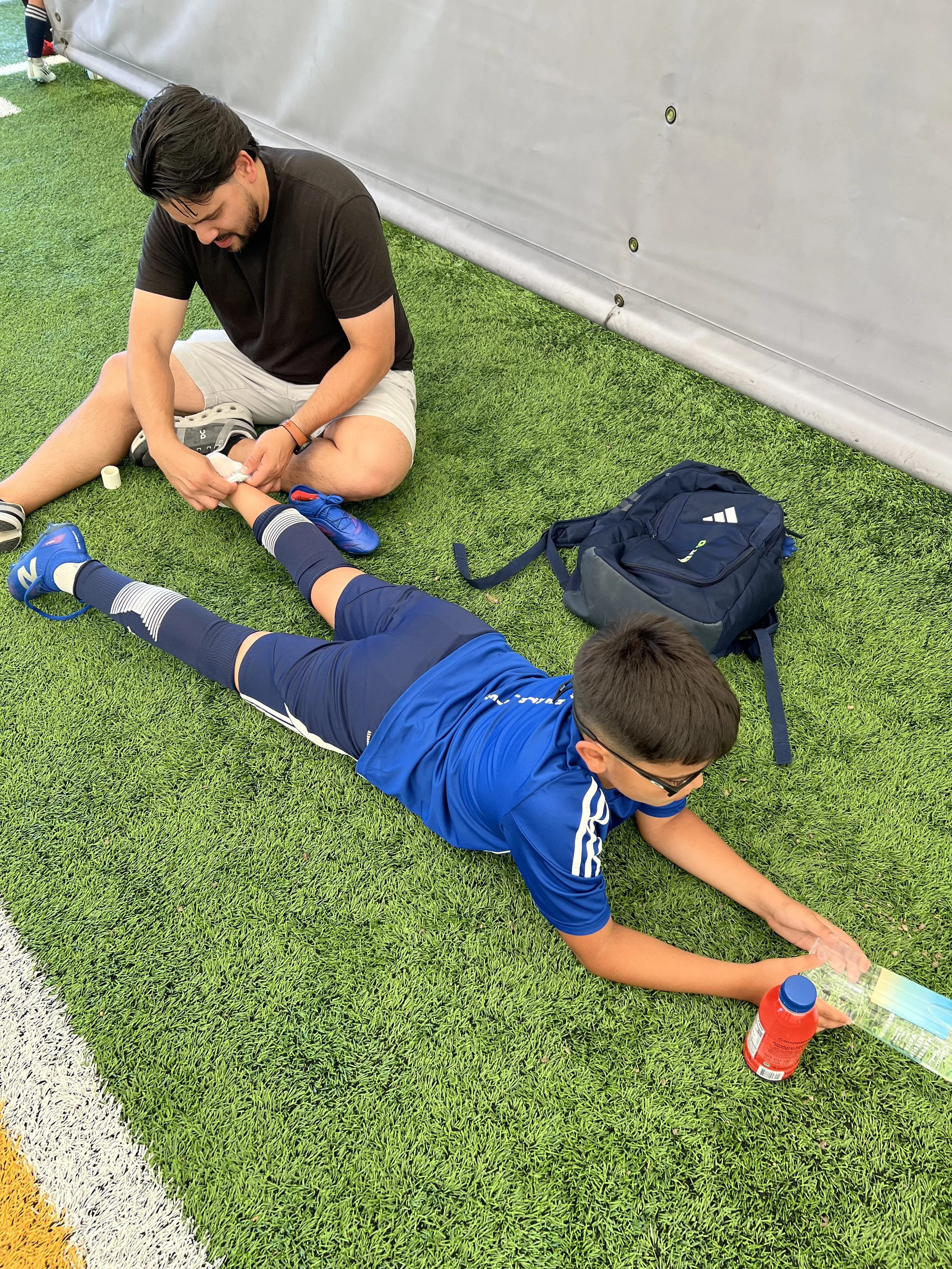 A young man in a black T-shirt and shorts is sitting on the artificial grass beside a soccer field, wrapping a medical bandage around a child's ankle. The child is lying on their stomach, reaching out with one arm to grab a colored bottle, possibly water or sports drink. There is a blue backpack on the ground nearby, and a white and yellow painted line marks the edge of the field.