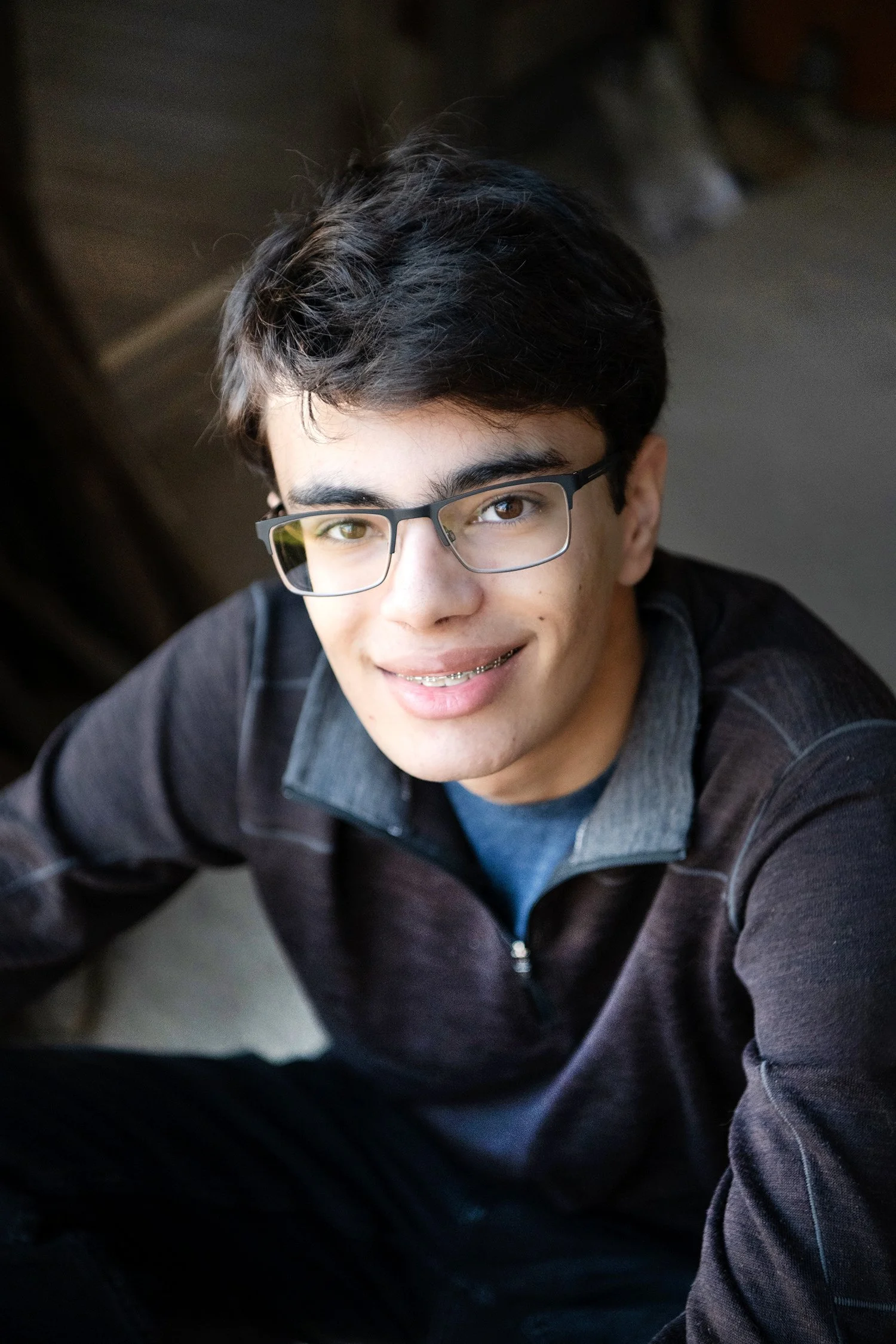 A young man with dark hair, glasses, and braces smiling at the camera, wearing a dark jacket with a grey collar, sitting indoors.