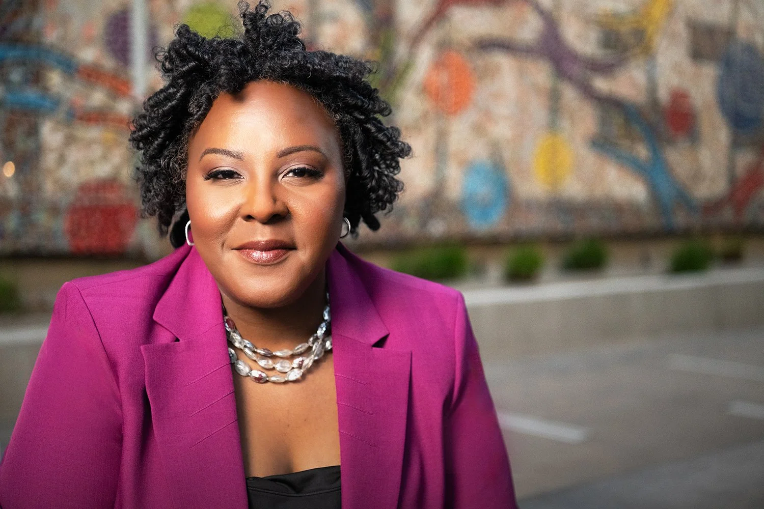 A woman with short curly black hair, wearing a pink blazer, a black top, and a pearl necklace, standing outdoors in front of a colorful mural.