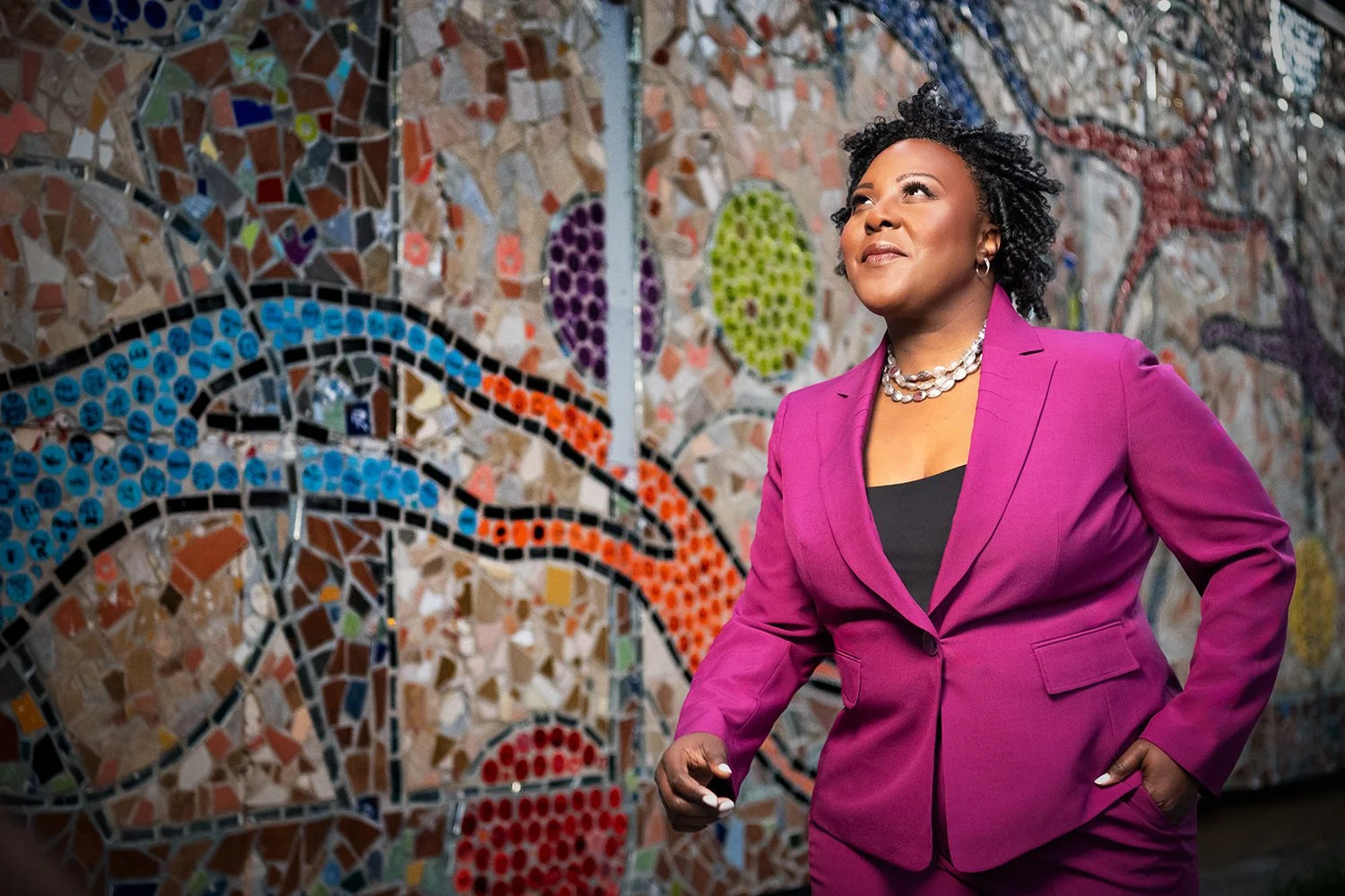 A woman wearing a magenta blazer and black top, standing in front of a colorful mosaic wall, looking upward.