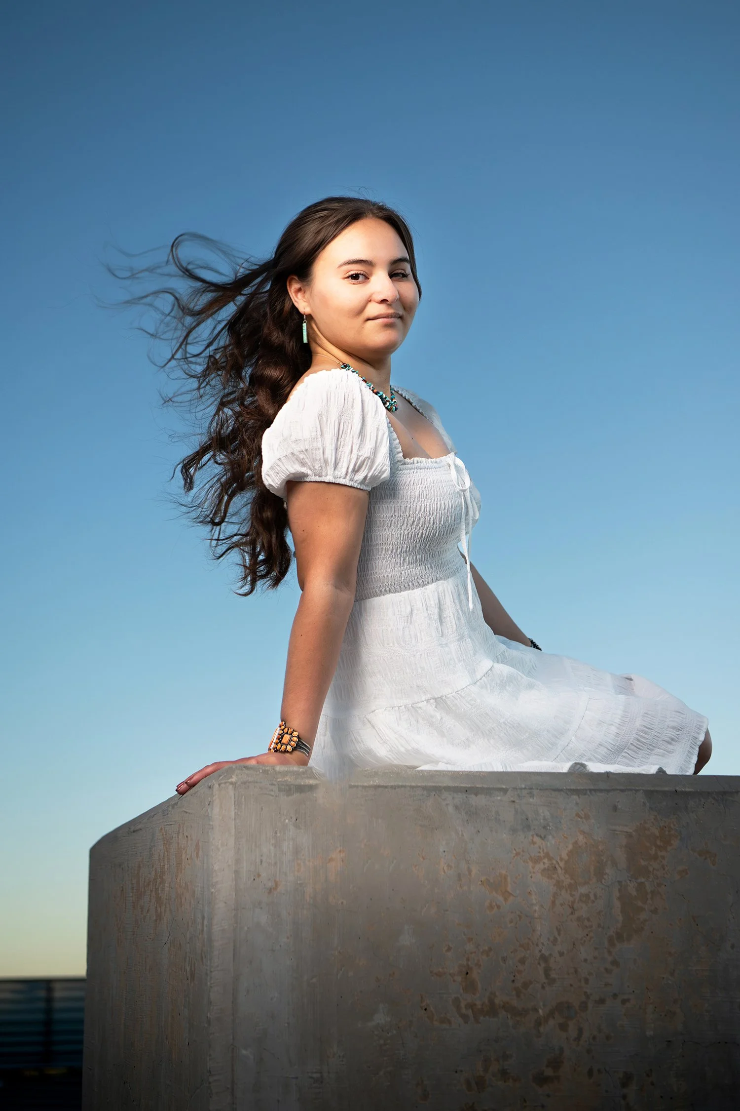 A woman with long dark hair sitting on a concrete ledge with a clear blue sky in the background, wearing a white dress, turquoise jewelry, and a large bracelet, looking at the camera with a calm expression during a senior portrait shoot in Tucson