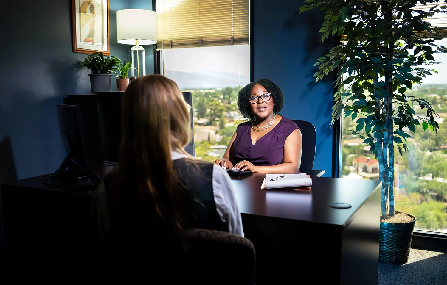 Two women engaged in a conversation in an office with large windows showing a city view, one woman is speaking, the other listens, with a computer monitor, plants, and framed art on the wall.