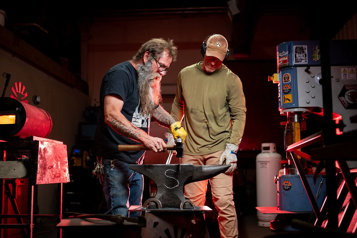 Two men working at a blacksmithing forge. The man on the left with a long gray beard and glasses is using a hammer, while the other man in a brown cap and headphones watches. The workspace is filled with tools and equipment.
