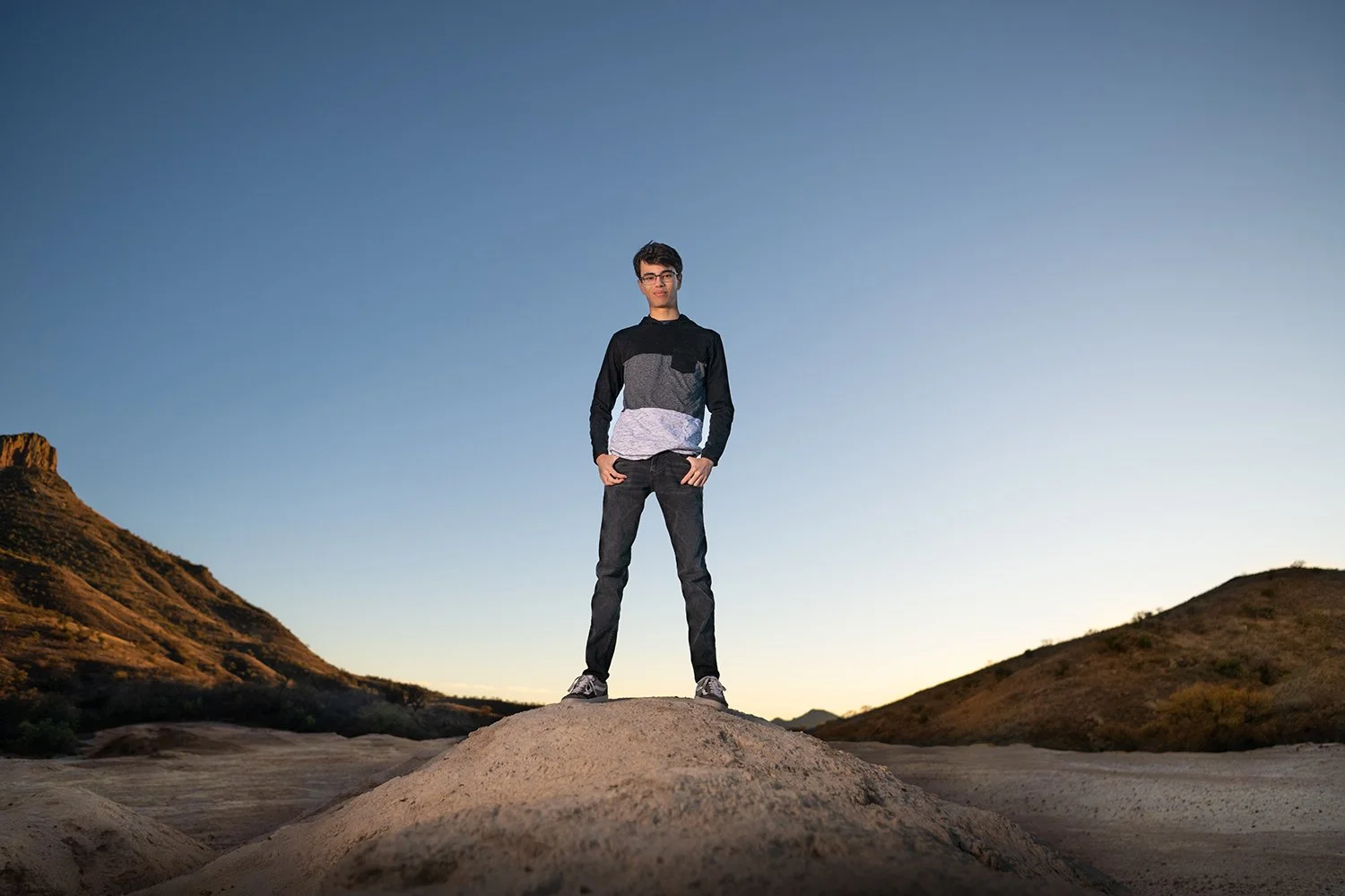 A high school senior standing on a large rock outdoors during sunset, with mountains or hills in the background and a clear sky for his senior portraits in Tucson