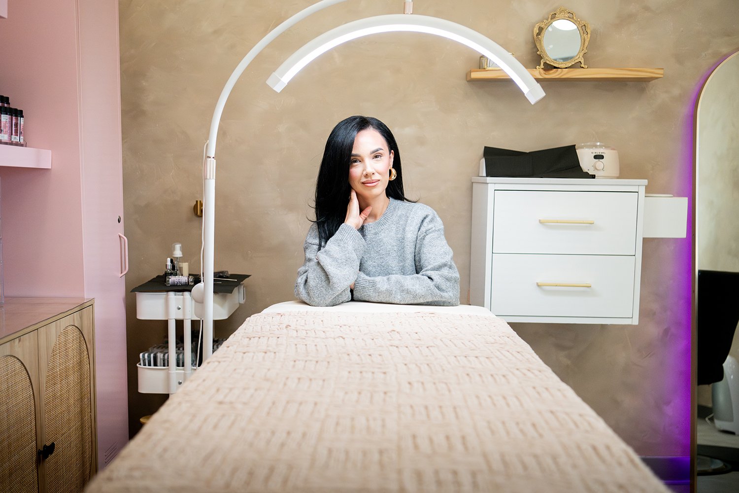 A woman with black hair and a gray sweater sitting at a massage table in a beauty or spa room, with a large mirror above her and various salon or spa equipment around her.