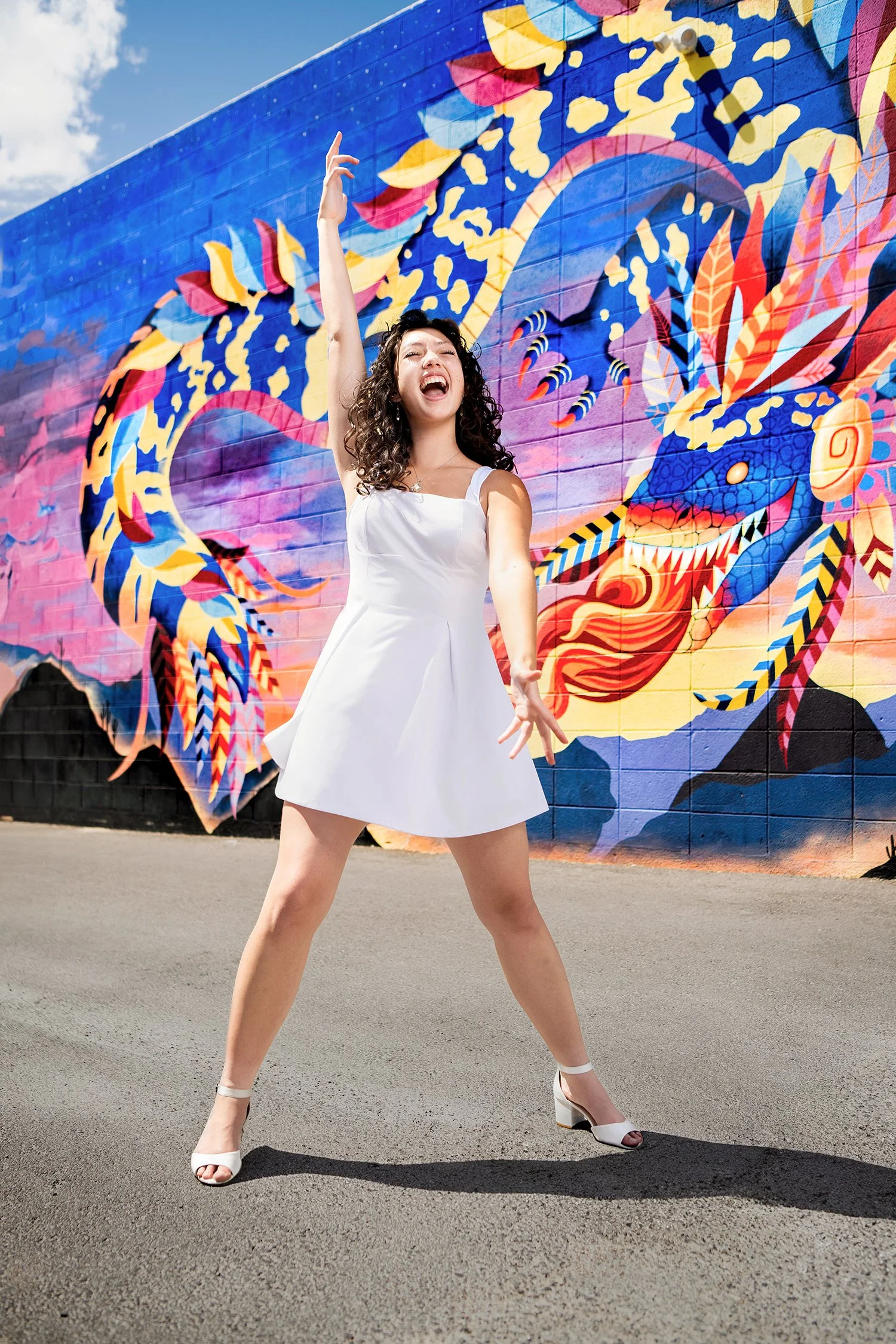 A woman in a white dress dancing in front of a colorful mural, with her right arm raised and her mouth open in a joyful expression.